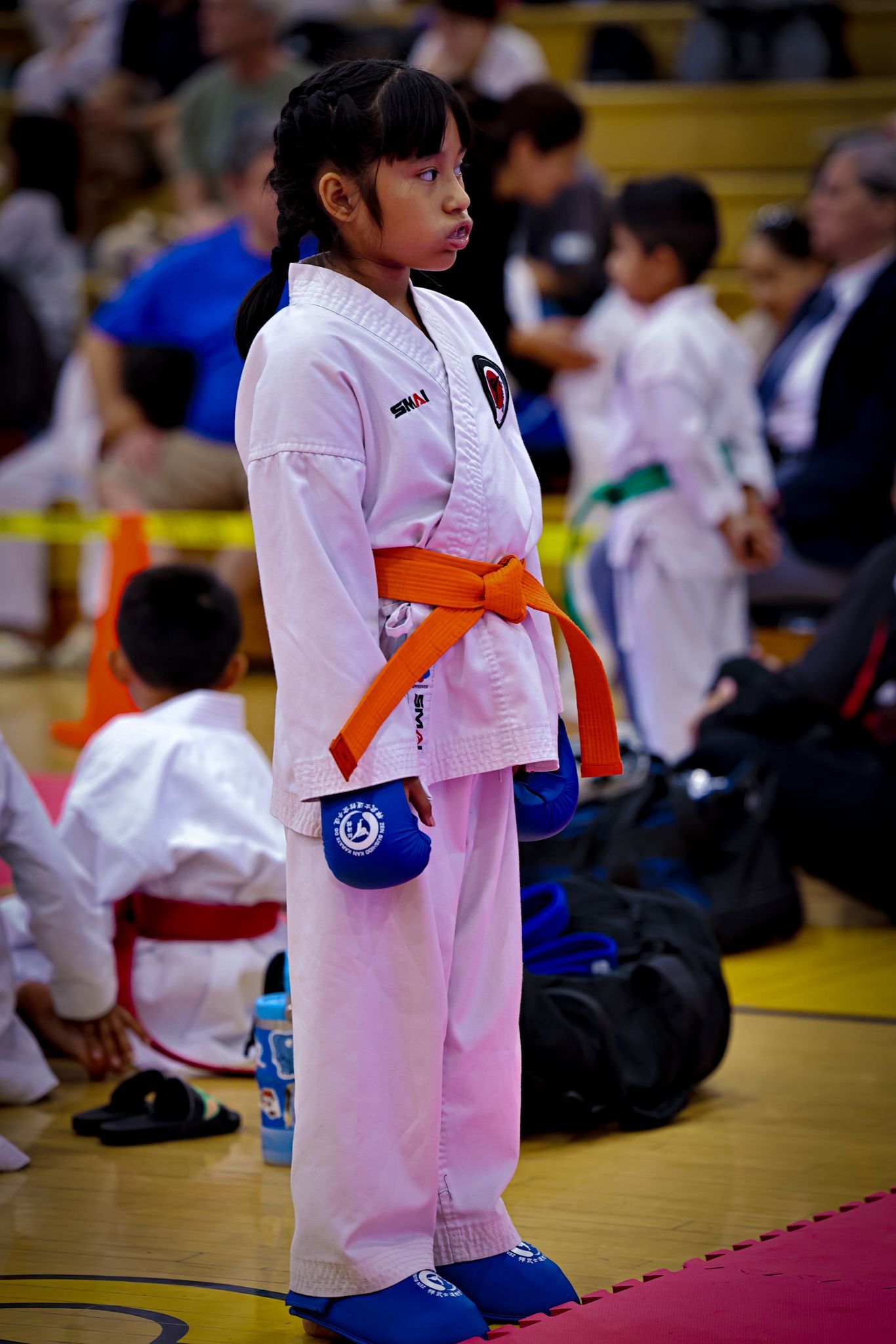 A martial artist in a white gi and orange belt stands on a gym floor, wearing blue gloves and foot pads.