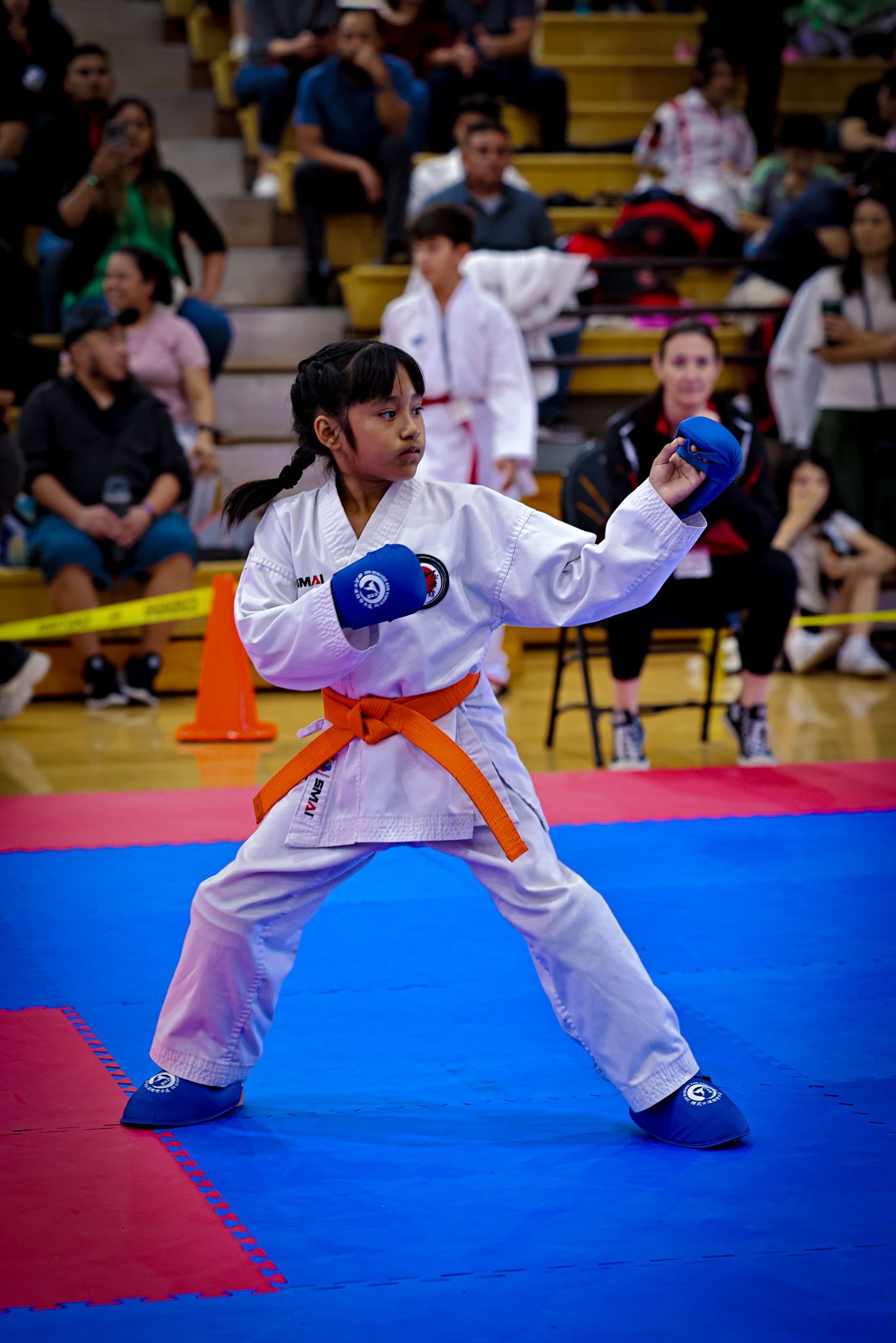 A person in a white karate gi and orange belt strikes a pose with blue sparring gloves in a gymnasium.