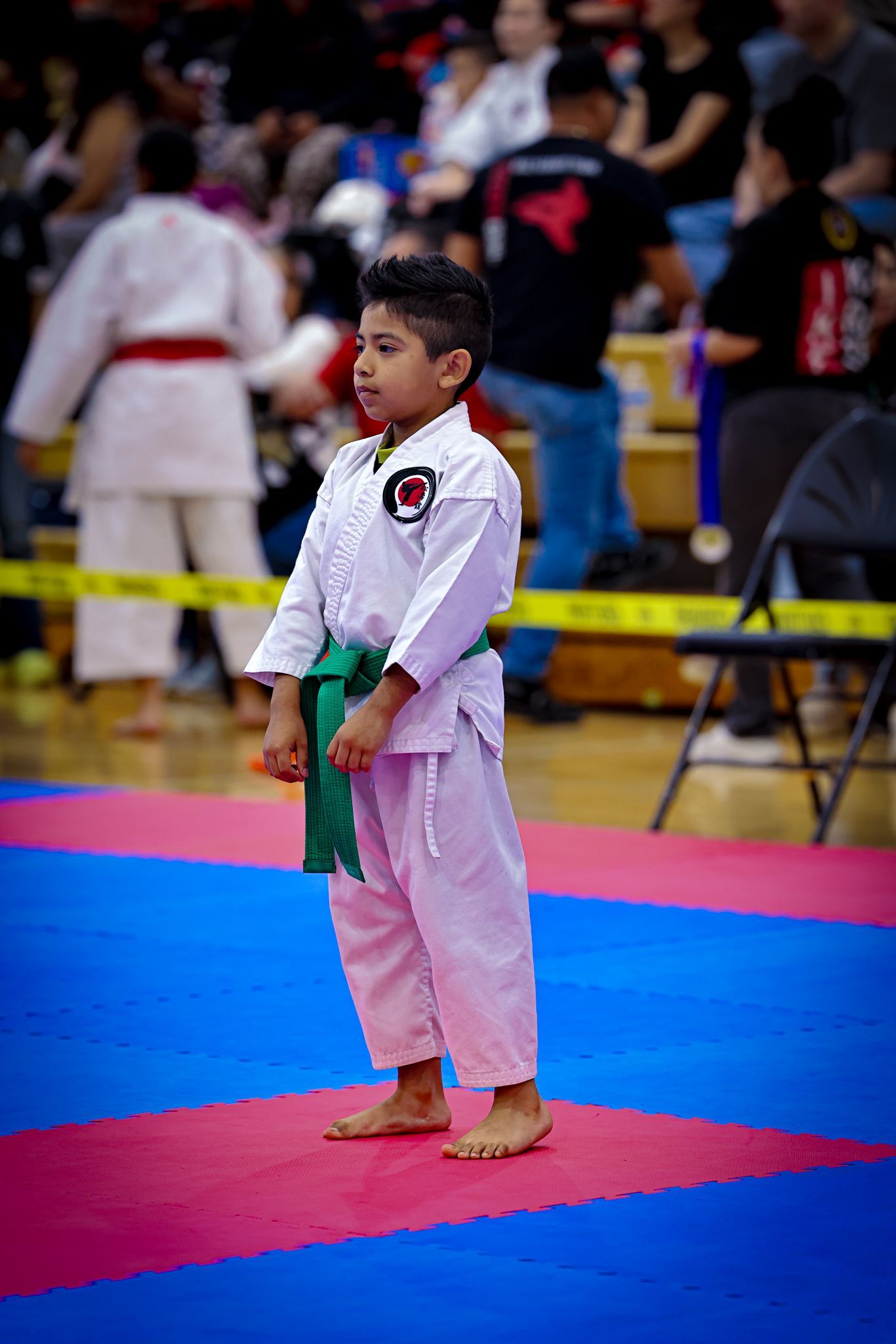 A young martial arts student with a green belt standing barefoot on a blue and pink mat at an indoor competition.