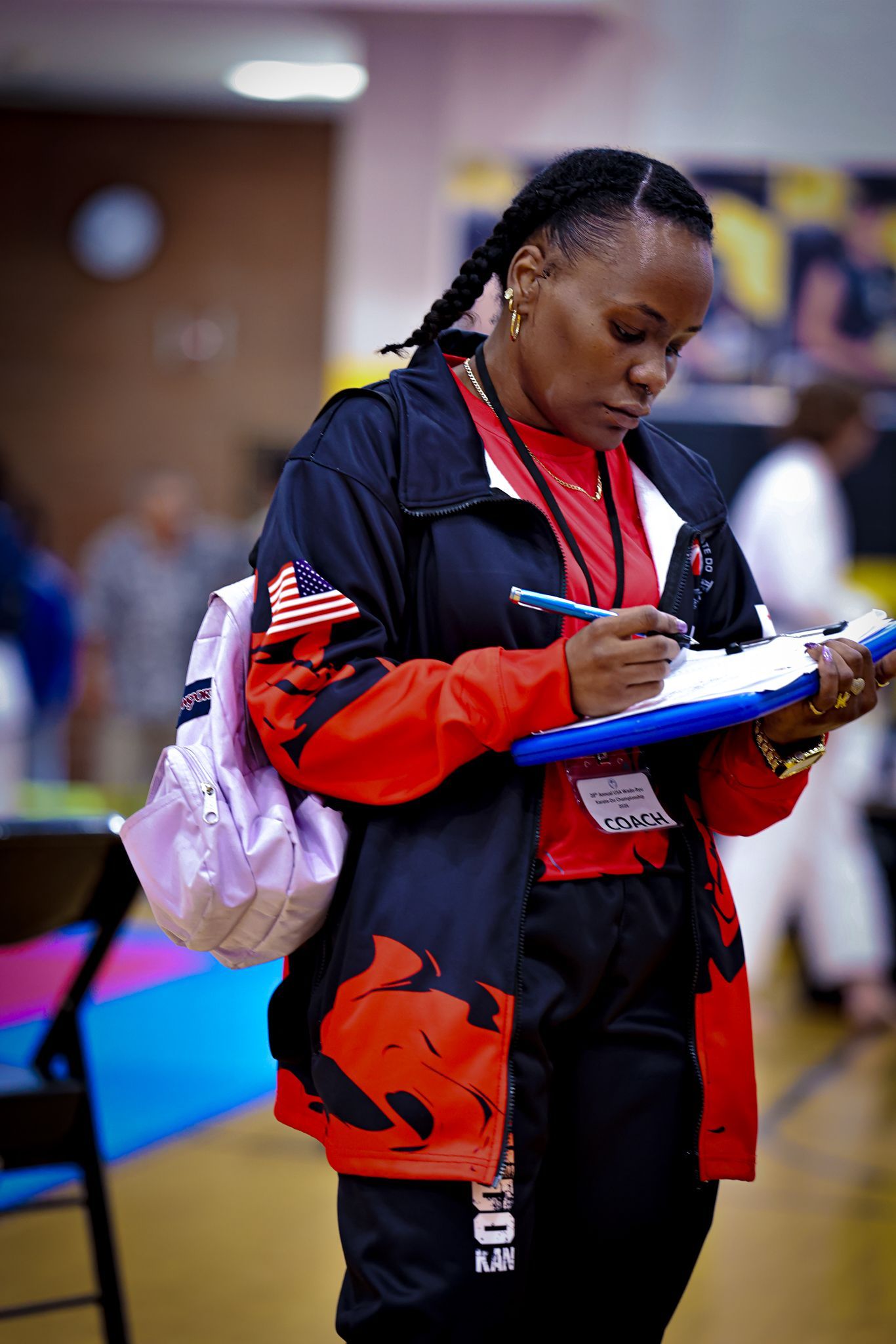 A woman wearing a black and red jacket and a light-colored backpack writes on a blue clipboard in a gym setting.
