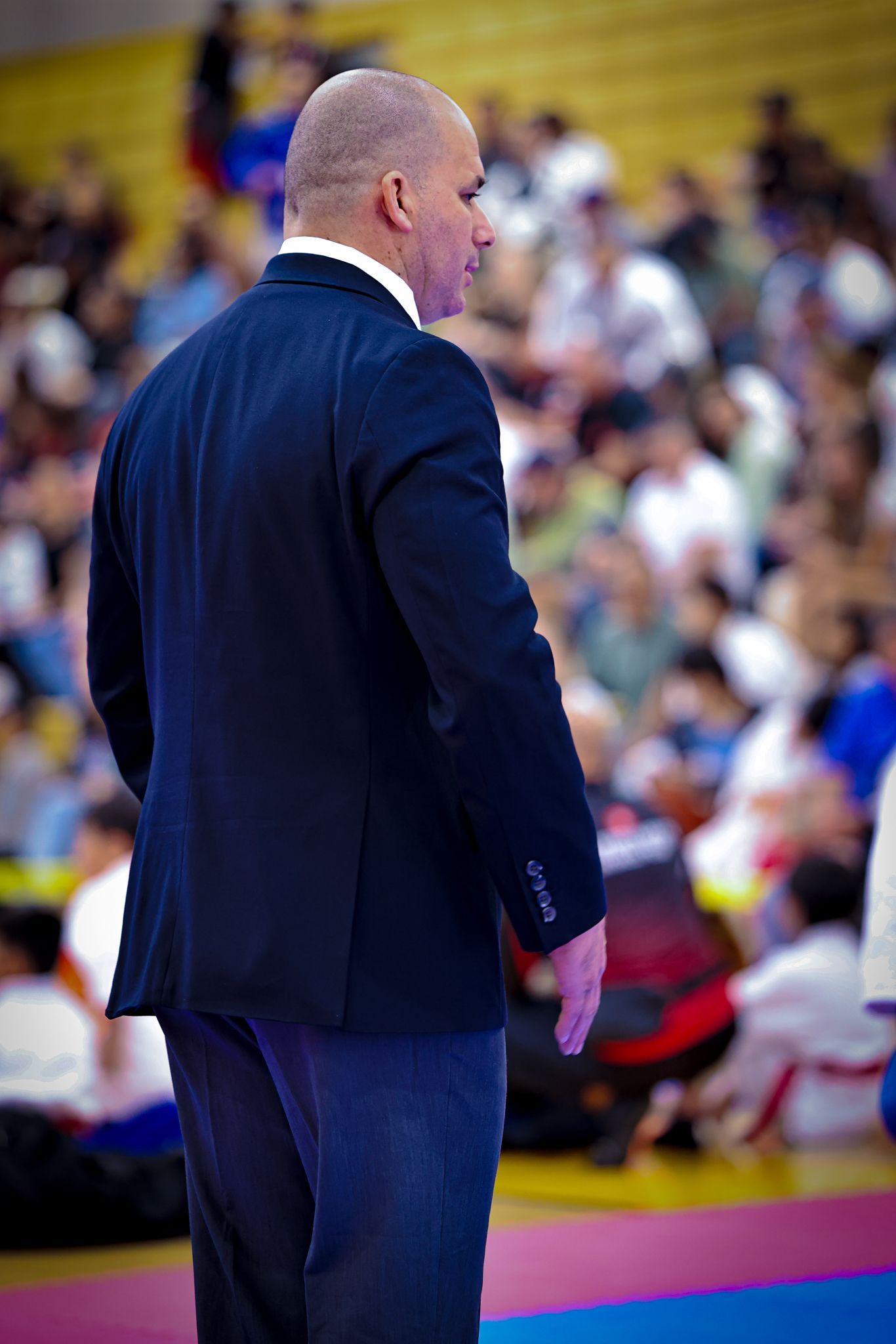 A coach in a dark blazer stands on the edge of a judo mat, watching a match in a crowded arena.