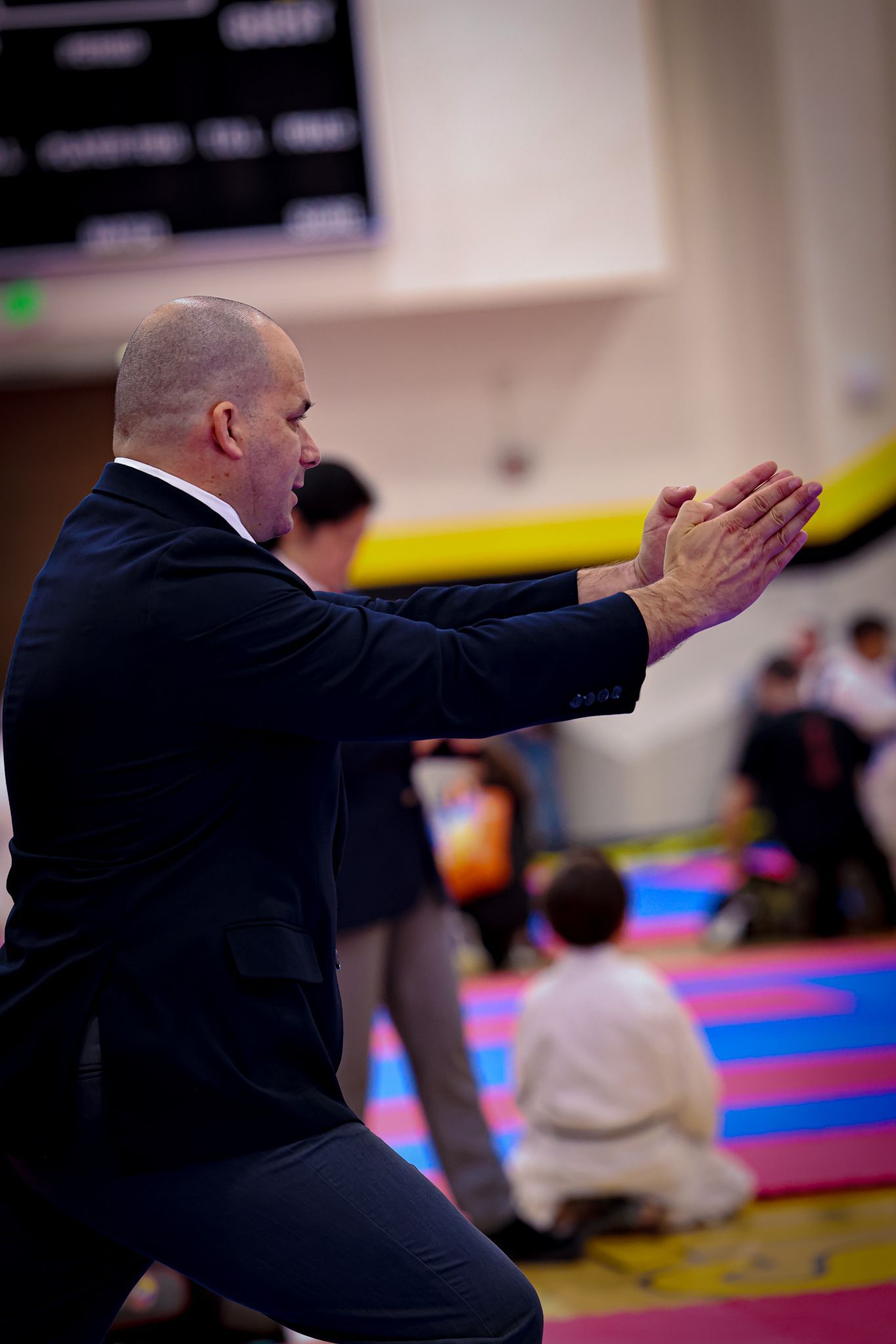 An instructor in a blazer performing a martial arts hand gesture in a gym, with a child in a uniform sitting nearby.