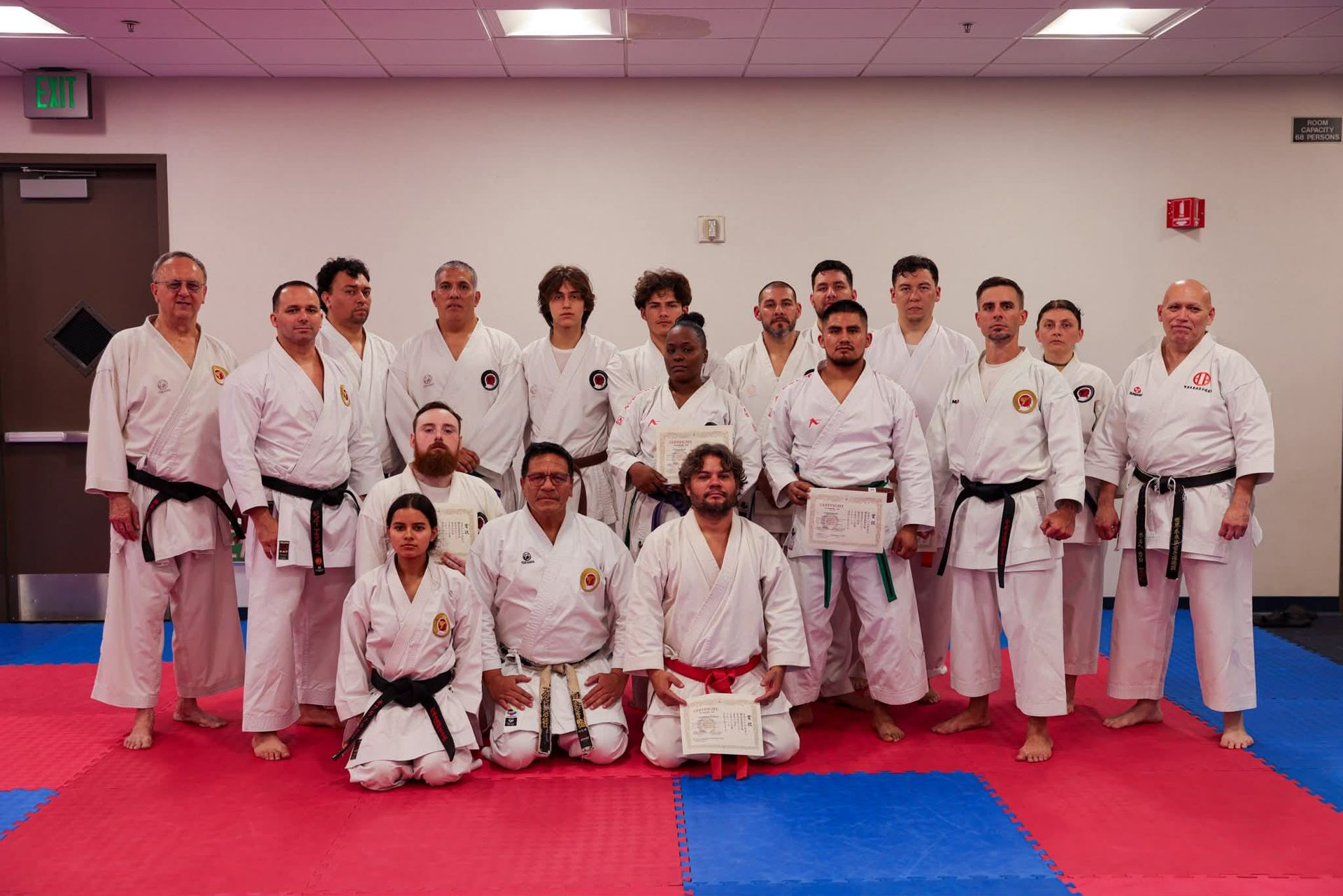 Group of karate practitioners in white uniforms, posing with certificates on a red and blue mat indoors.