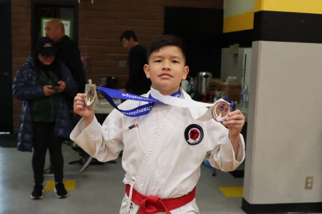 A young boy in a karate uniform is holding two medals around his neck.