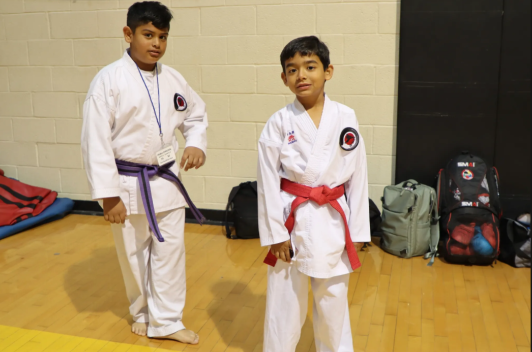 Two young boys in karate uniforms are standing next to each other on a wooden floor.