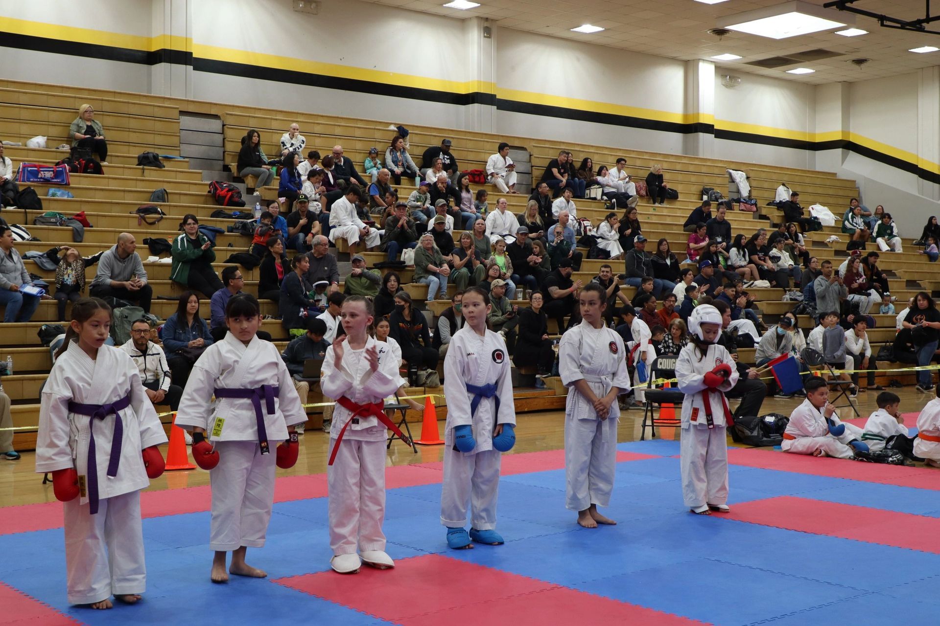 A group of children are standing on a karate mat in front of a crowd.
