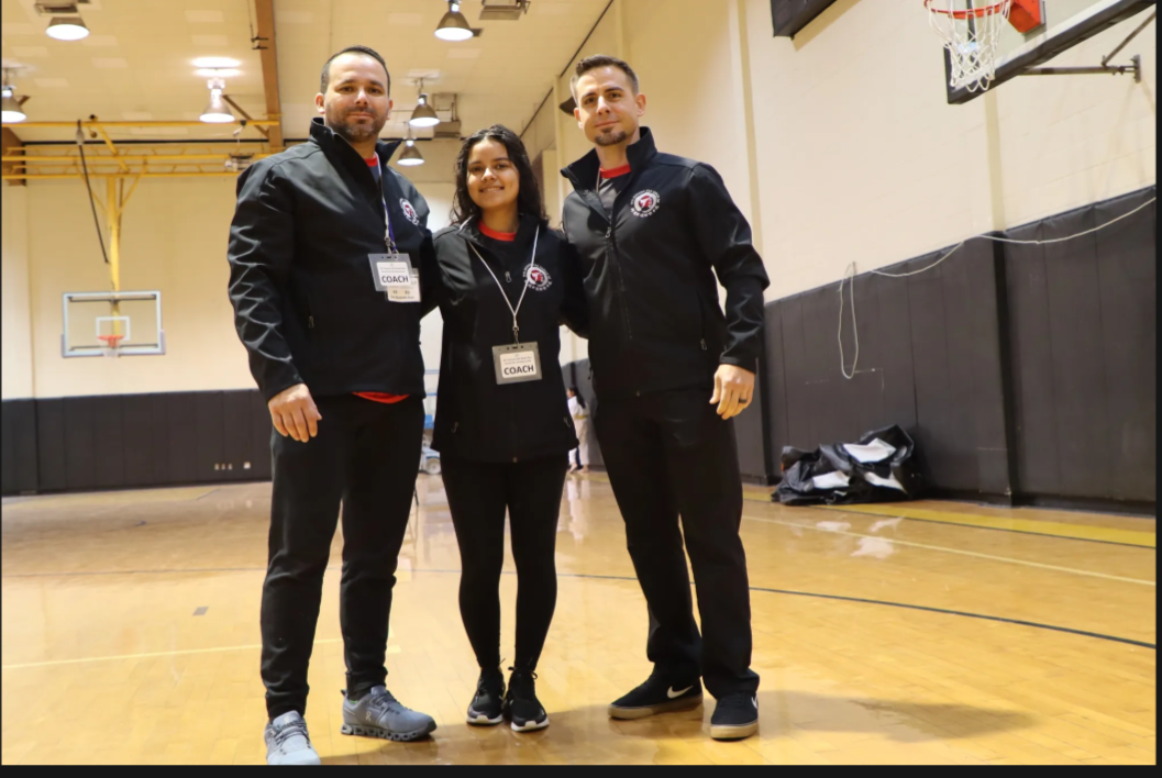 Three people are posing for a picture in a gym with a basketball hoop in the background.