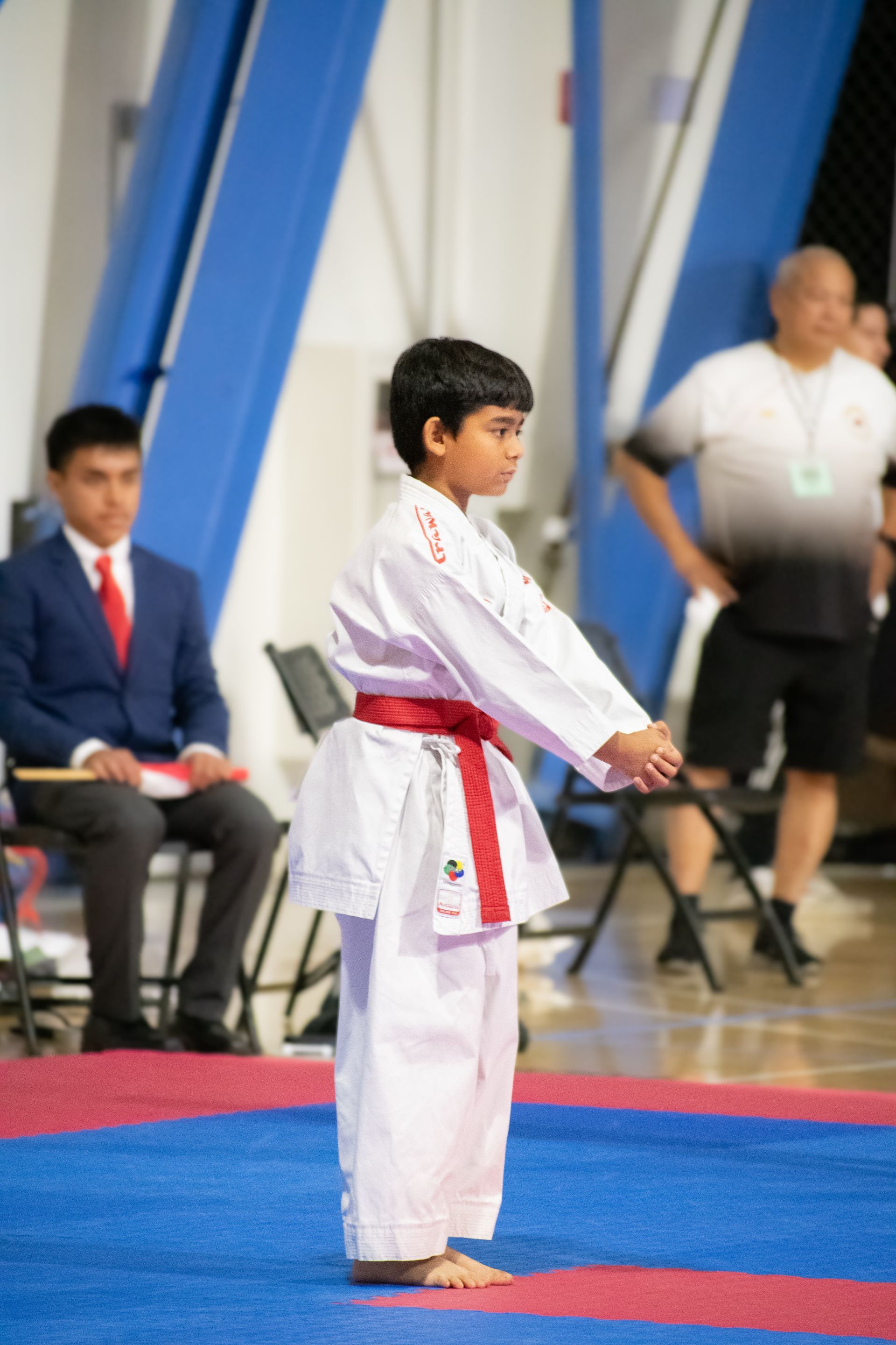 Boy in white karate uniform with red belt on blue mat. Officials watch in background.