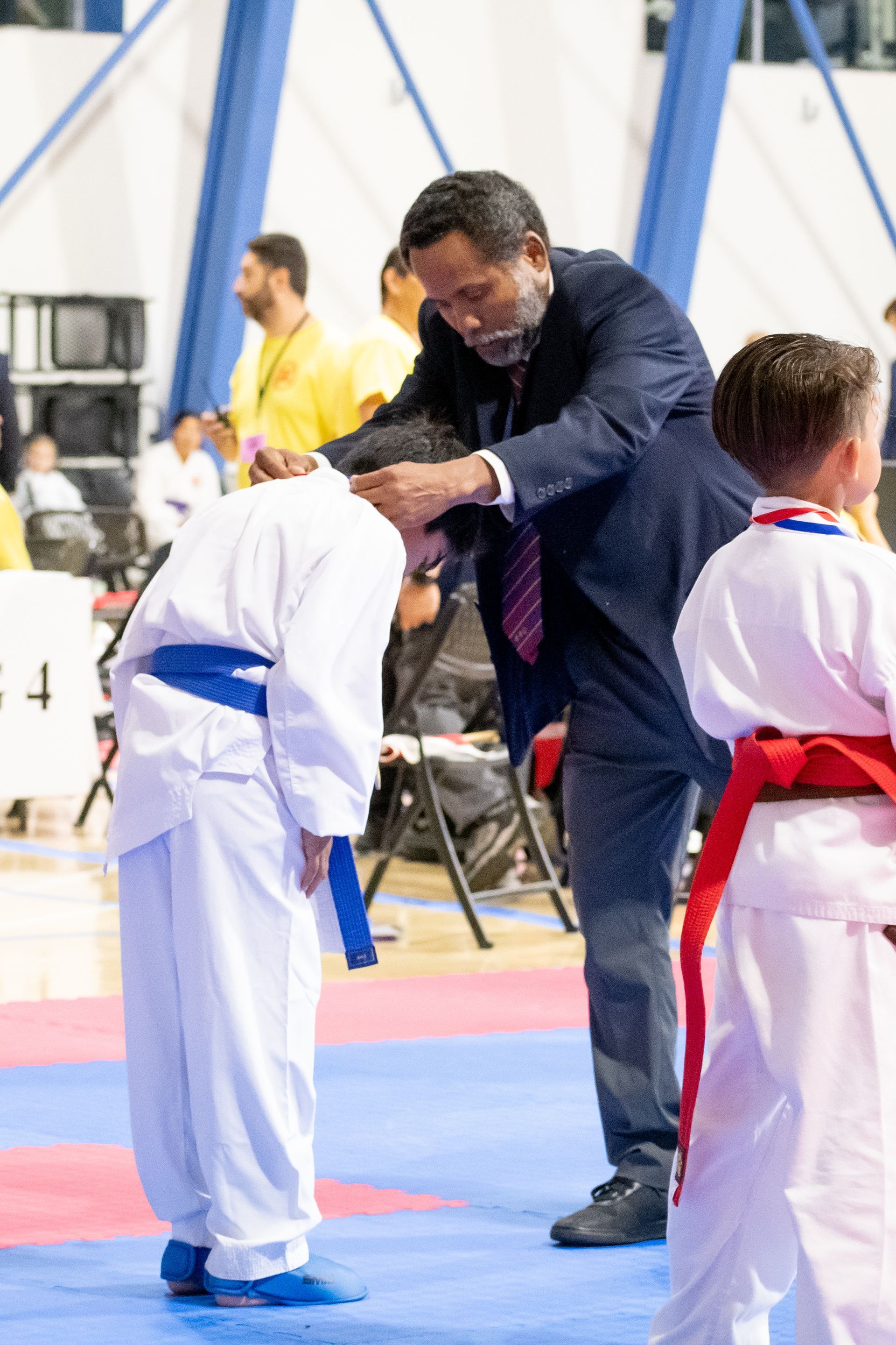 Man in suit awarding medal to a boy in karate uniform, bowing. Indoor sports setting.