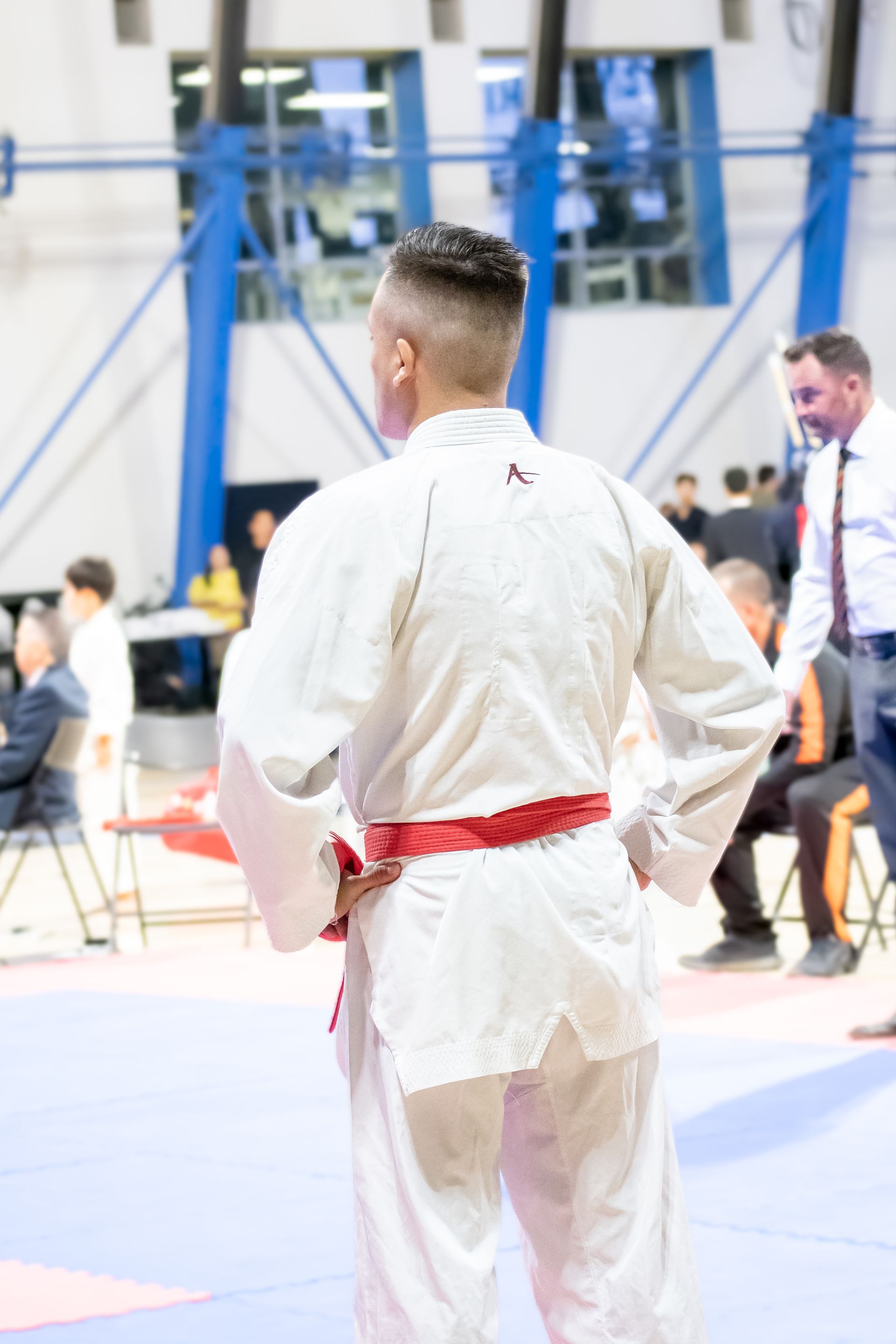 Martial artist in white uniform with red belt, back to camera, hands on hips in arena.