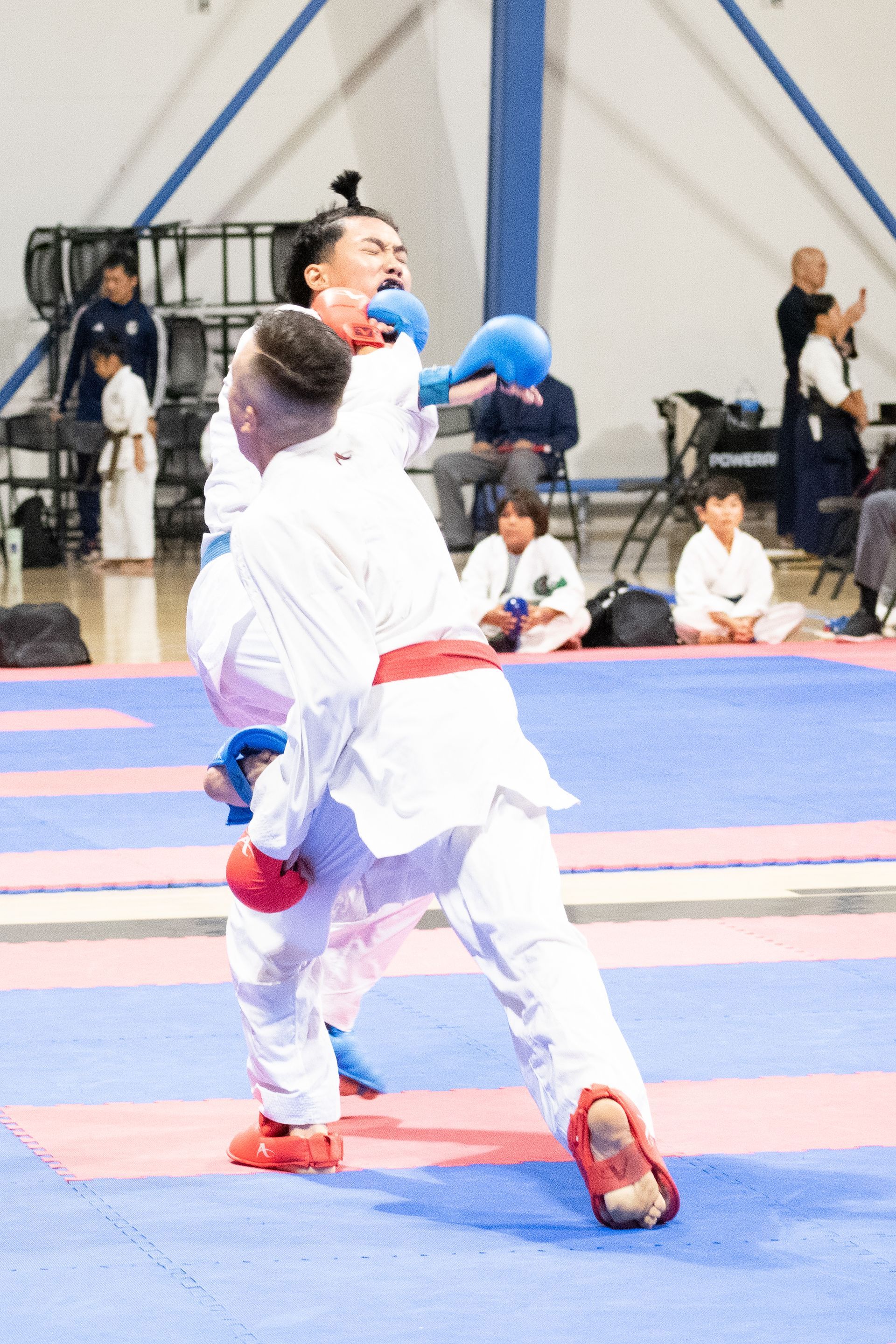Two karate fighters in white uniforms, one blocking a punch in a gymnasium setting.