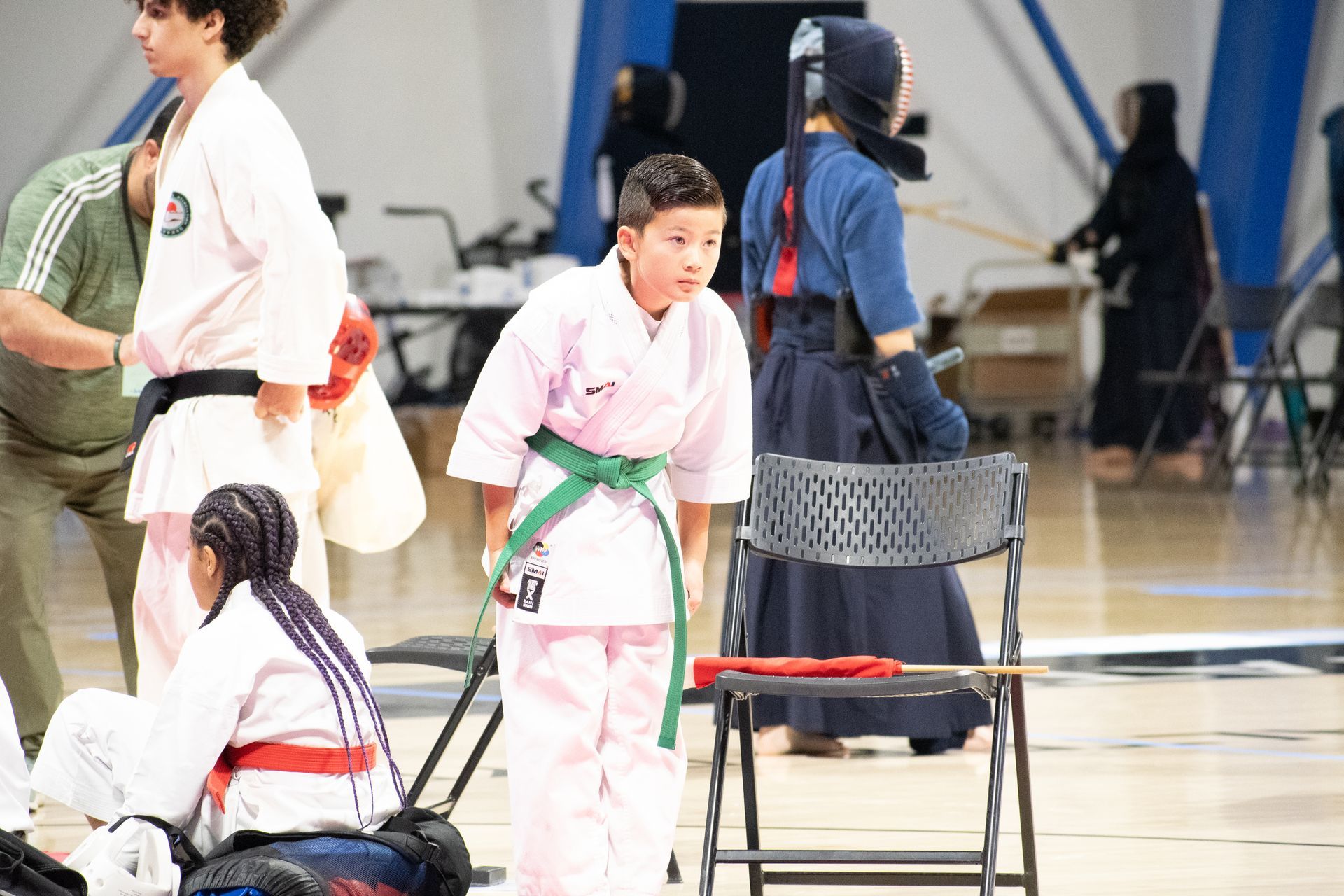 Boy in pink karate uniform with green belt, bowing in a gym, others in martial arts uniforms.