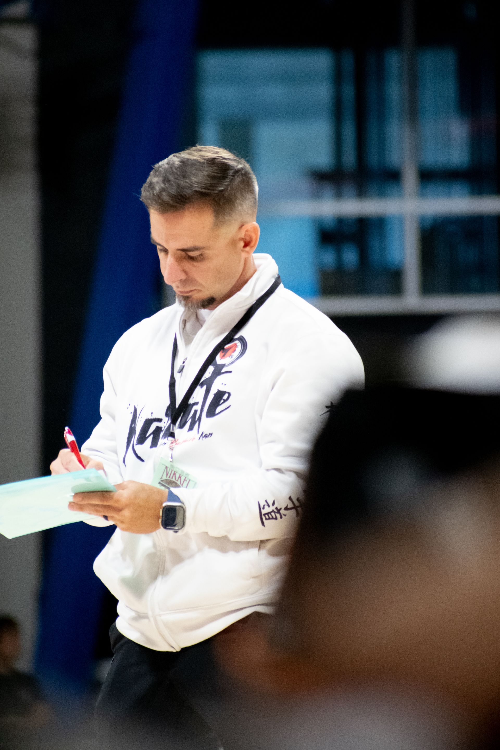 Man in white jacket taking notes on a clipboard in a basketball gym.