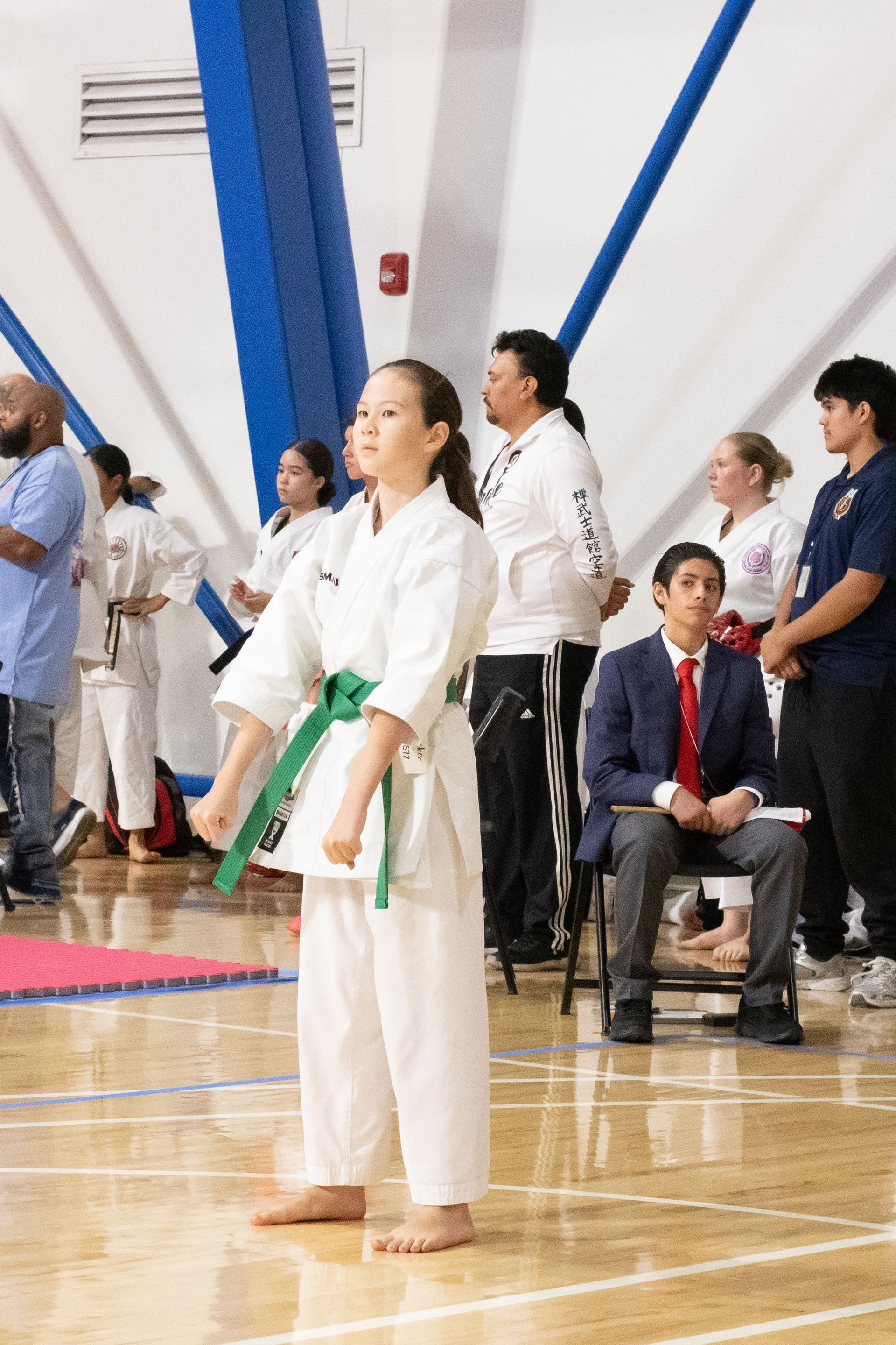 Girl in karate uniform, green belt, stands ready at tournament, gym setting.
