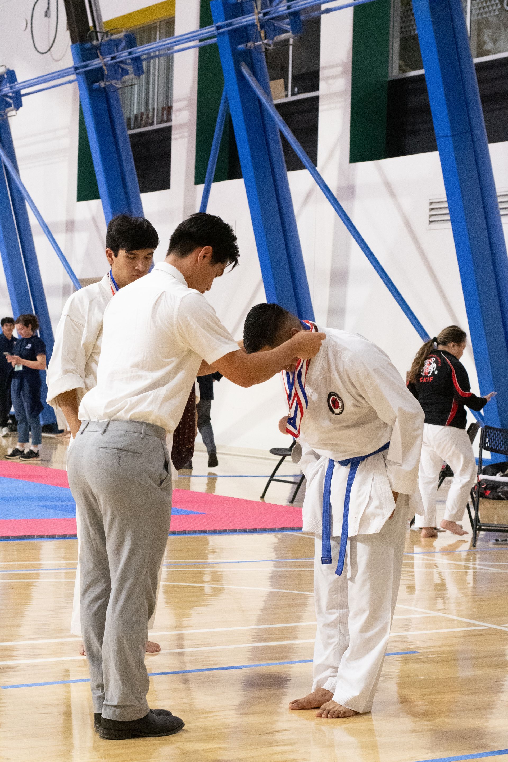 A karate competitor in white gi bows as a man places a medal around his neck in a gymnasium.