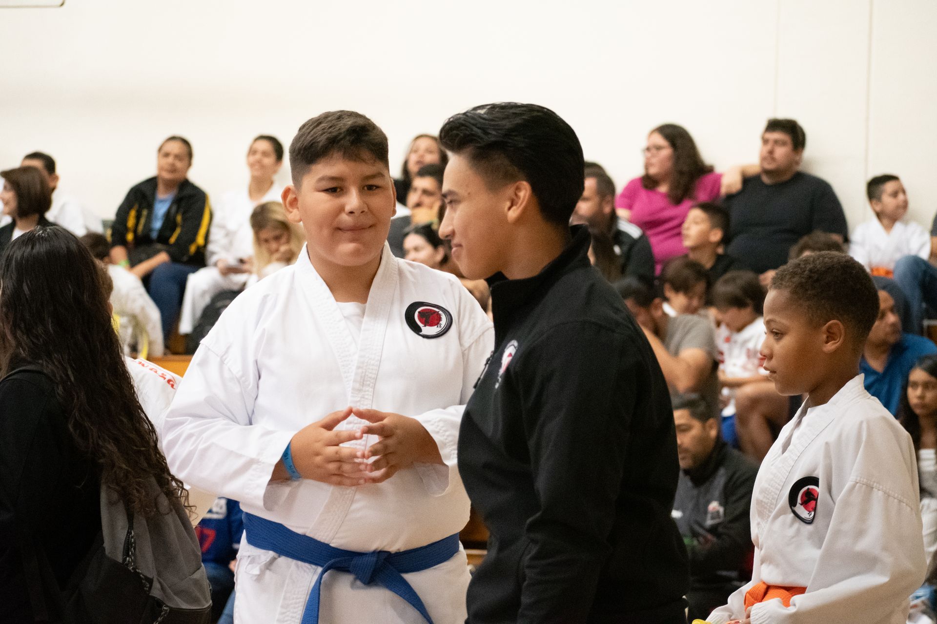 Two boys in karate uniforms are standing next to each other in front of a crowd.
