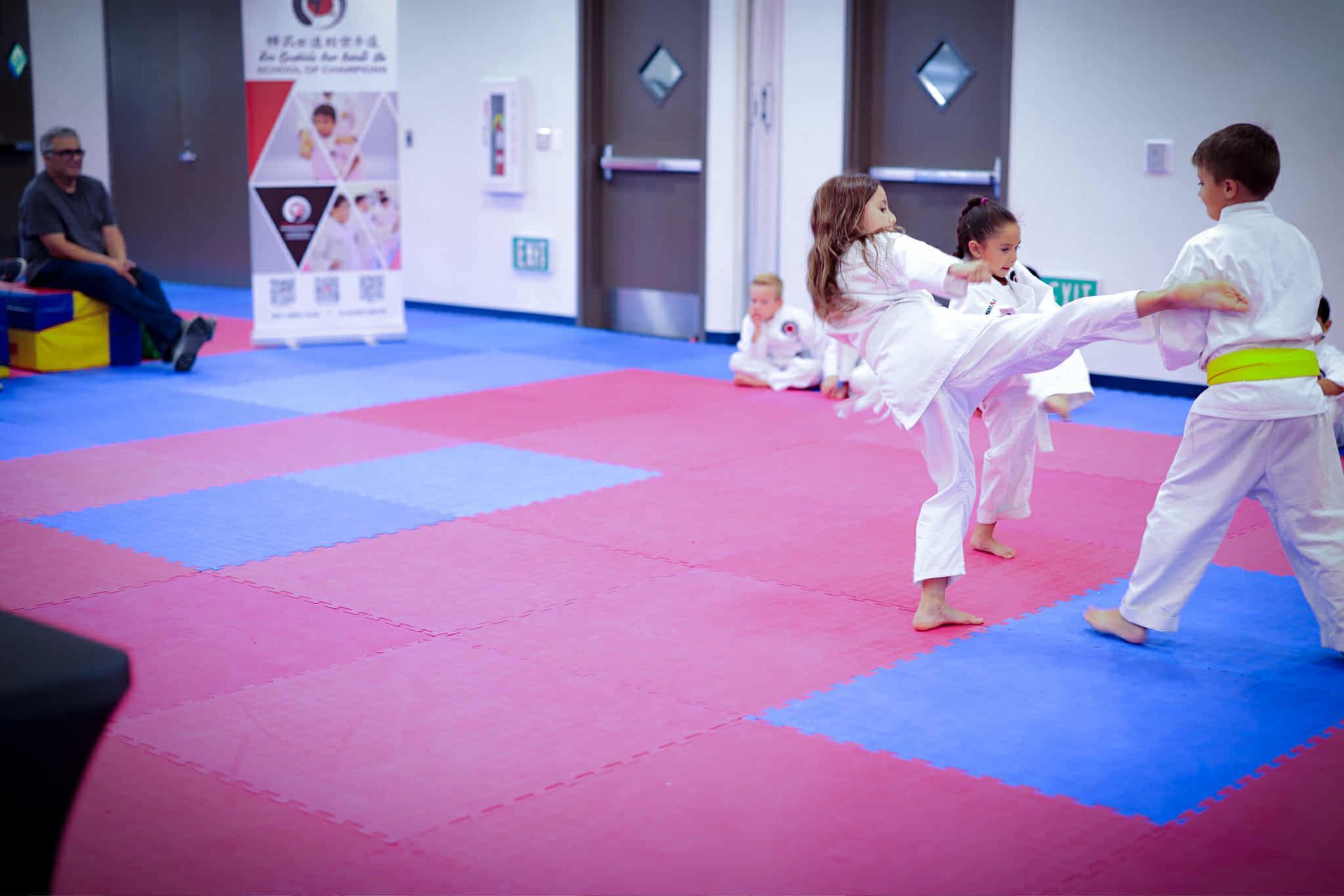 Children in white karate uniforms practicing kicks on a blue and red mat.