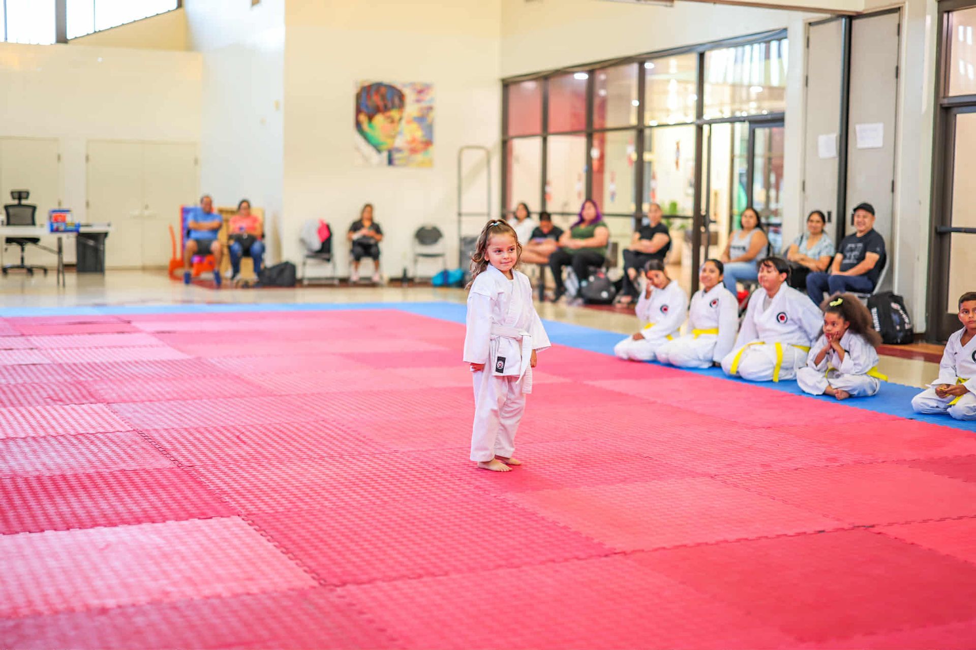 Young girl in karate uniform stands on a red and blue mat, facing a seated group; indoor gym setting.