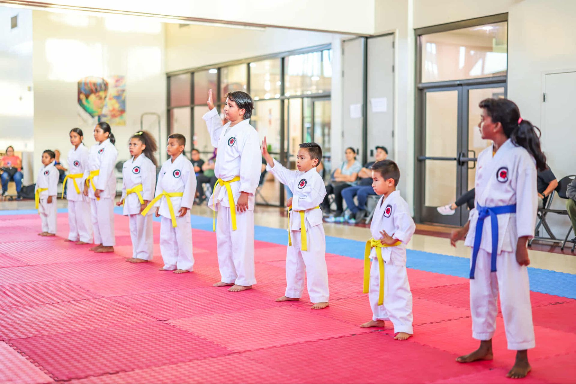 Children in karate uniforms, lined up on red mats, practicing in a dojo.