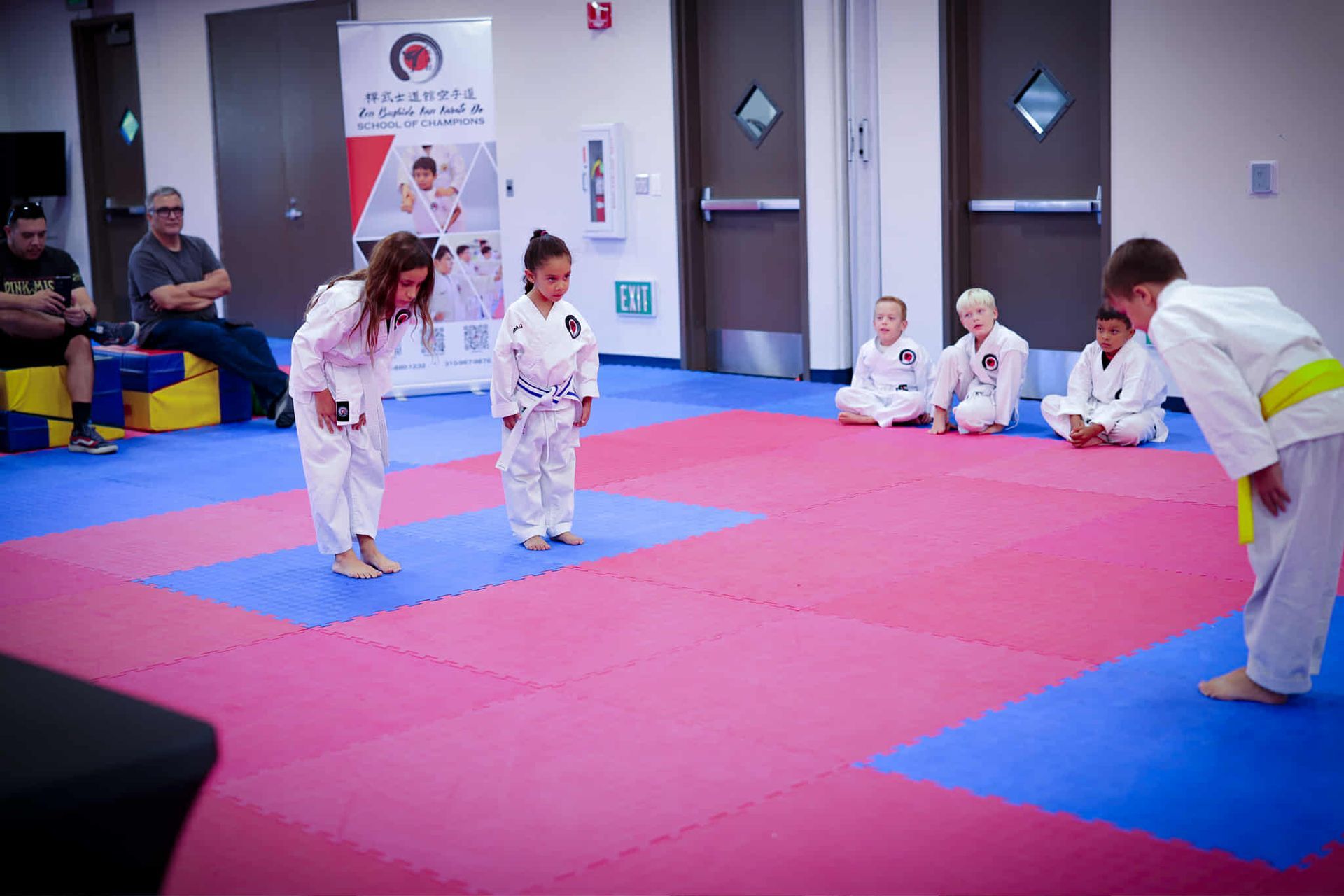 Kids in white karate uniforms bowing on a red and blue mat in a dojo.