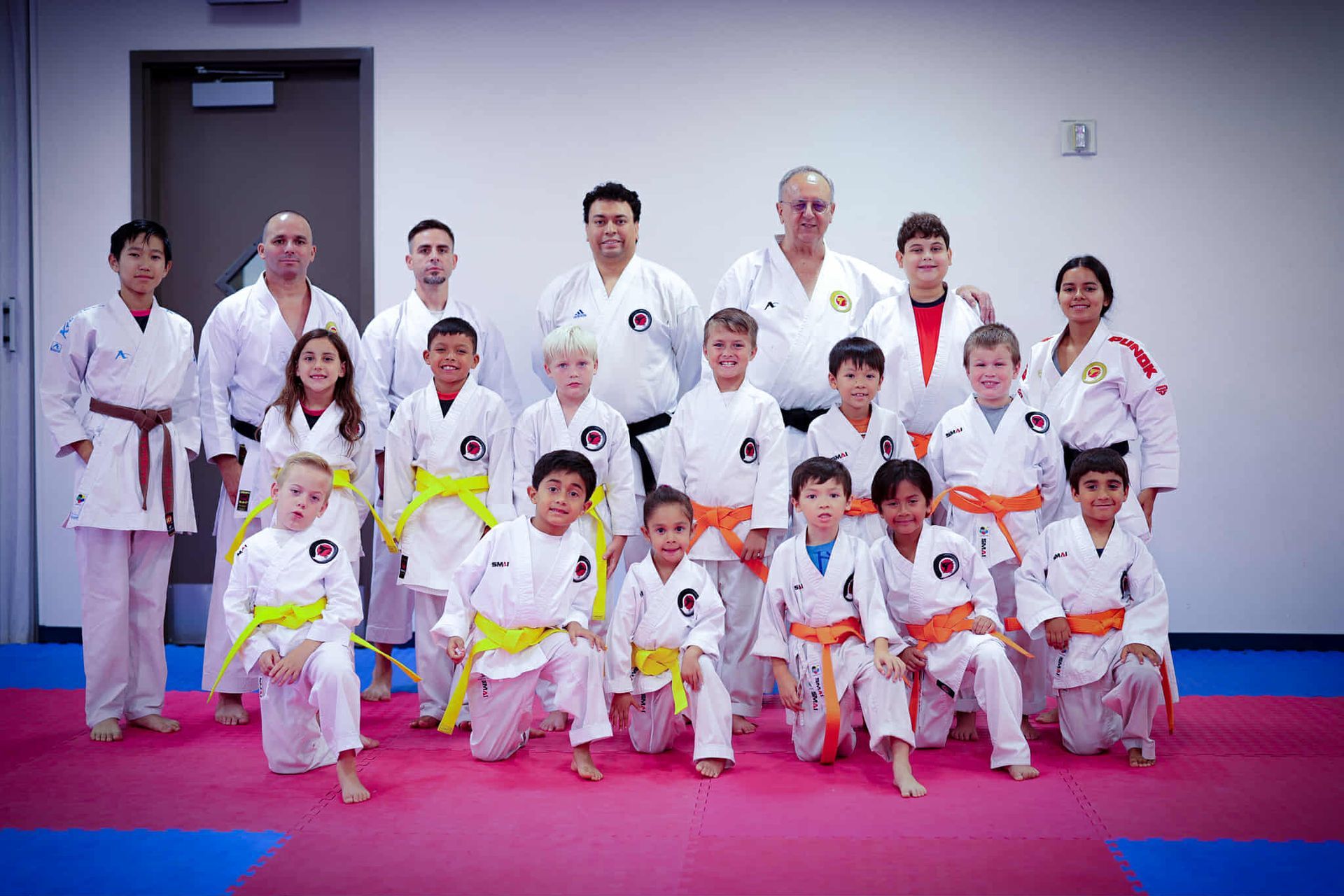 Karate class group photo: diverse students & instructors in white gis, varying belts, posed on a blue/red mat.