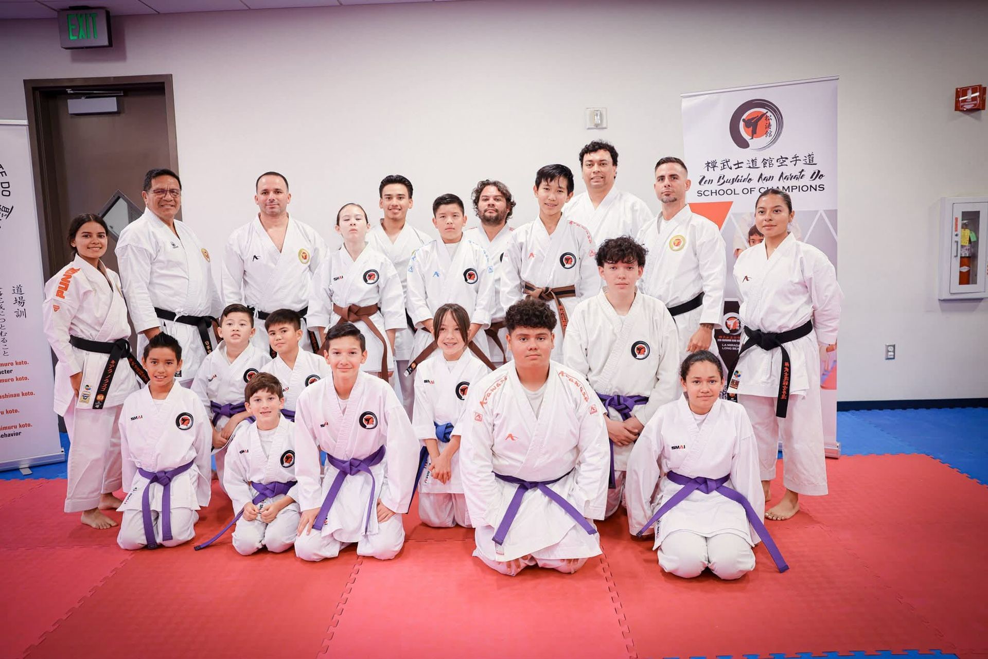 Group of karate practitioners in white uniforms, kneeling and standing on a red and blue mat, posing for a photo.
