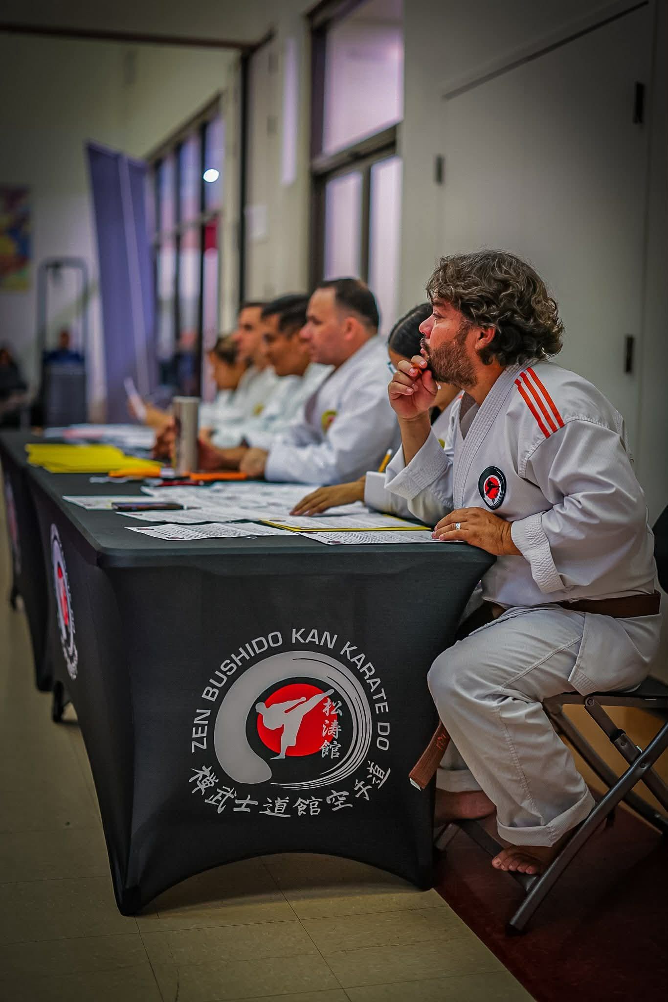 People in karate uniforms sitting at a table with the organization's logo, possibly judging a competition.