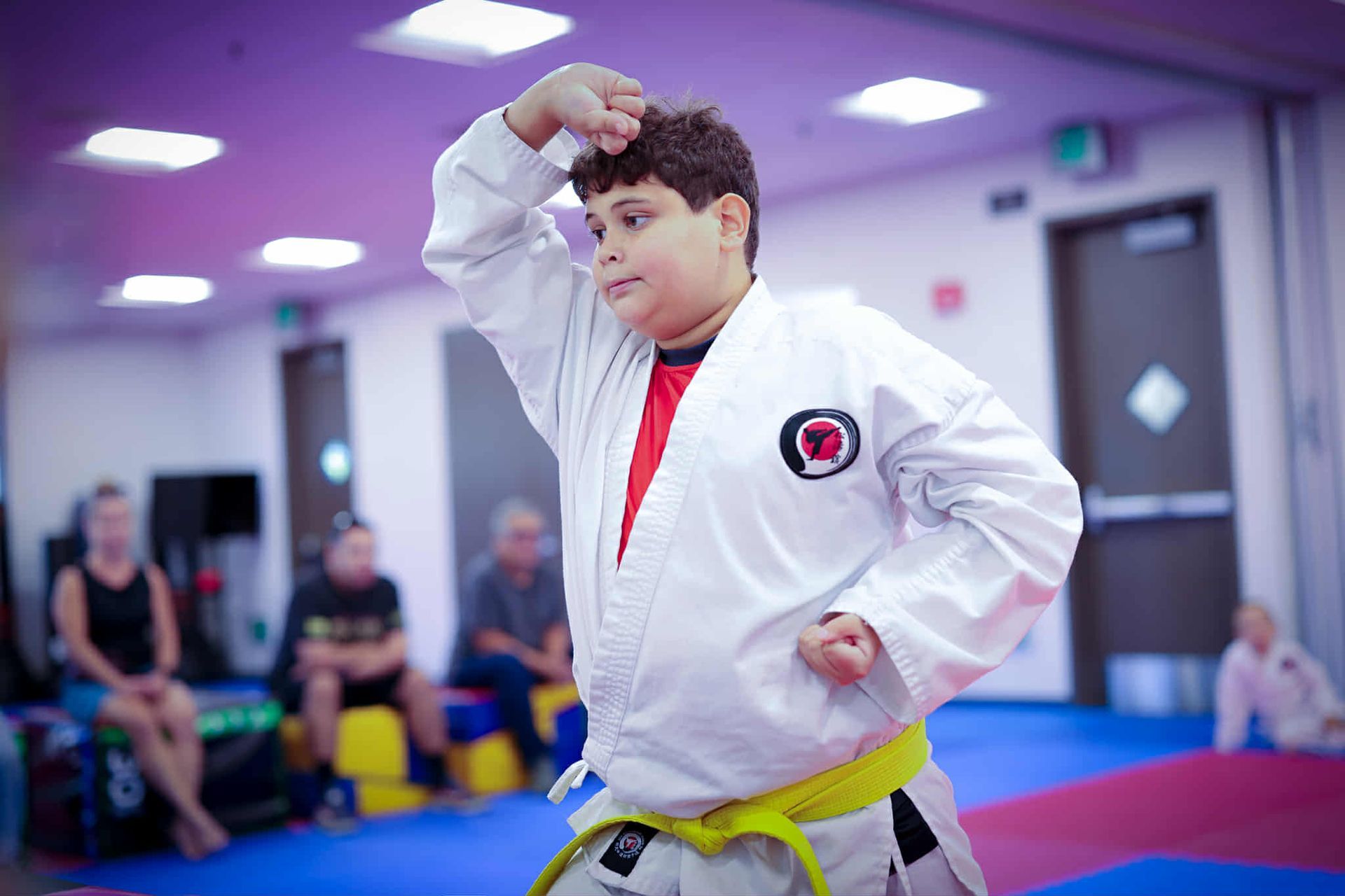 Young boy in karate uniform practices a move in a dojo. He has a yellow belt, focused expression.