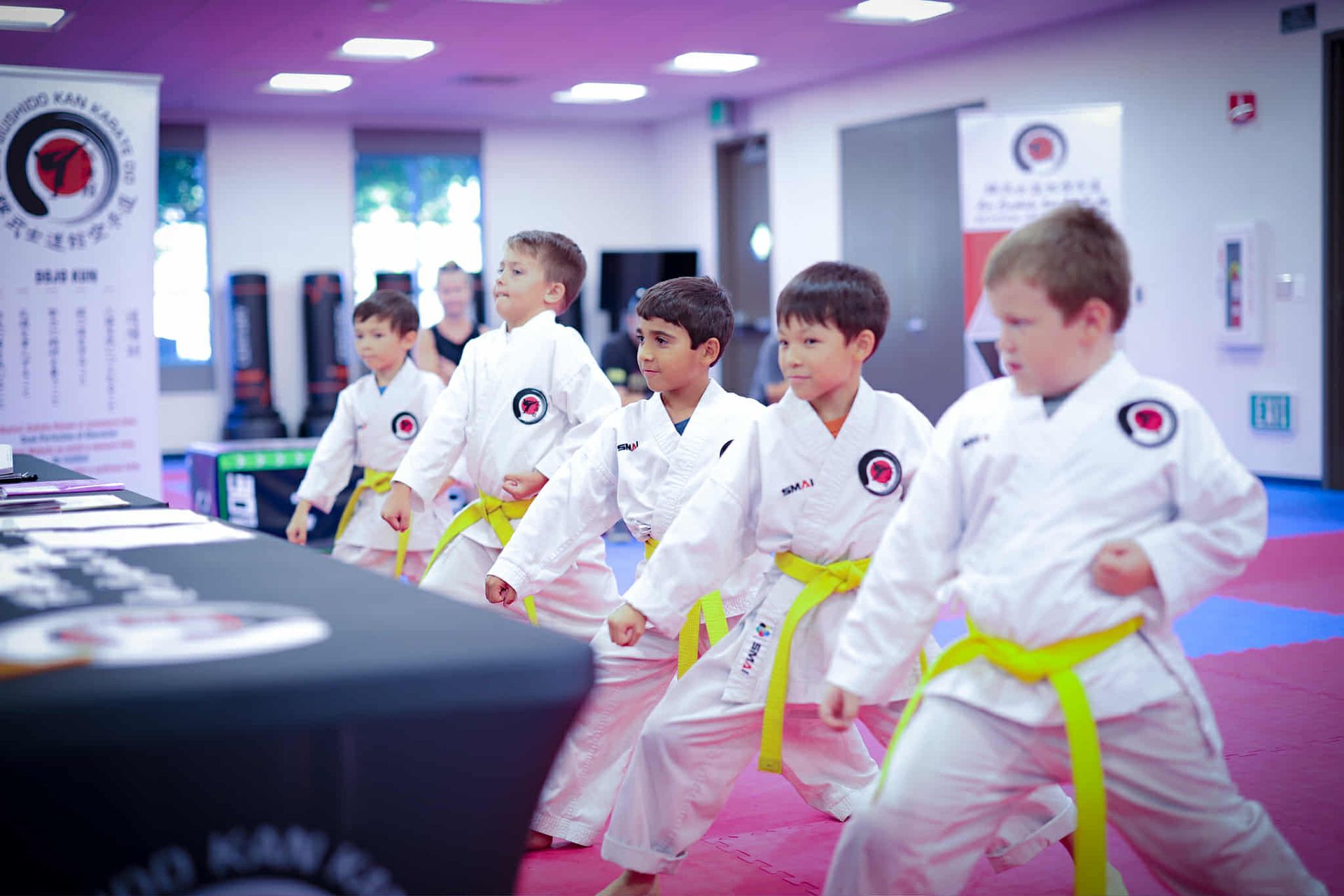 Children in white karate uniforms with yellow belts practice martial arts in a brightly lit room.