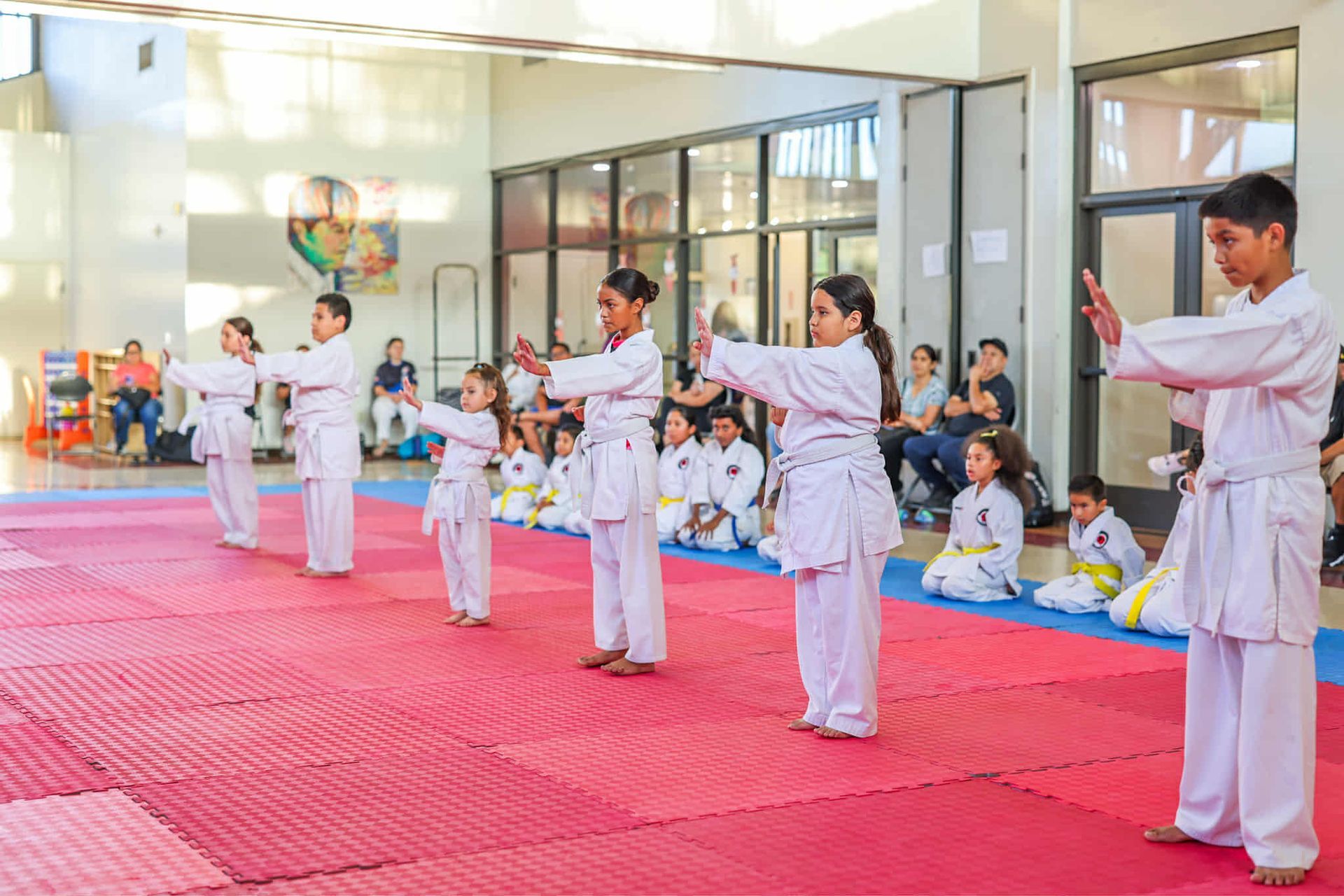 Children in white karate uniforms practicing a kata on red mats inside a bright gym.