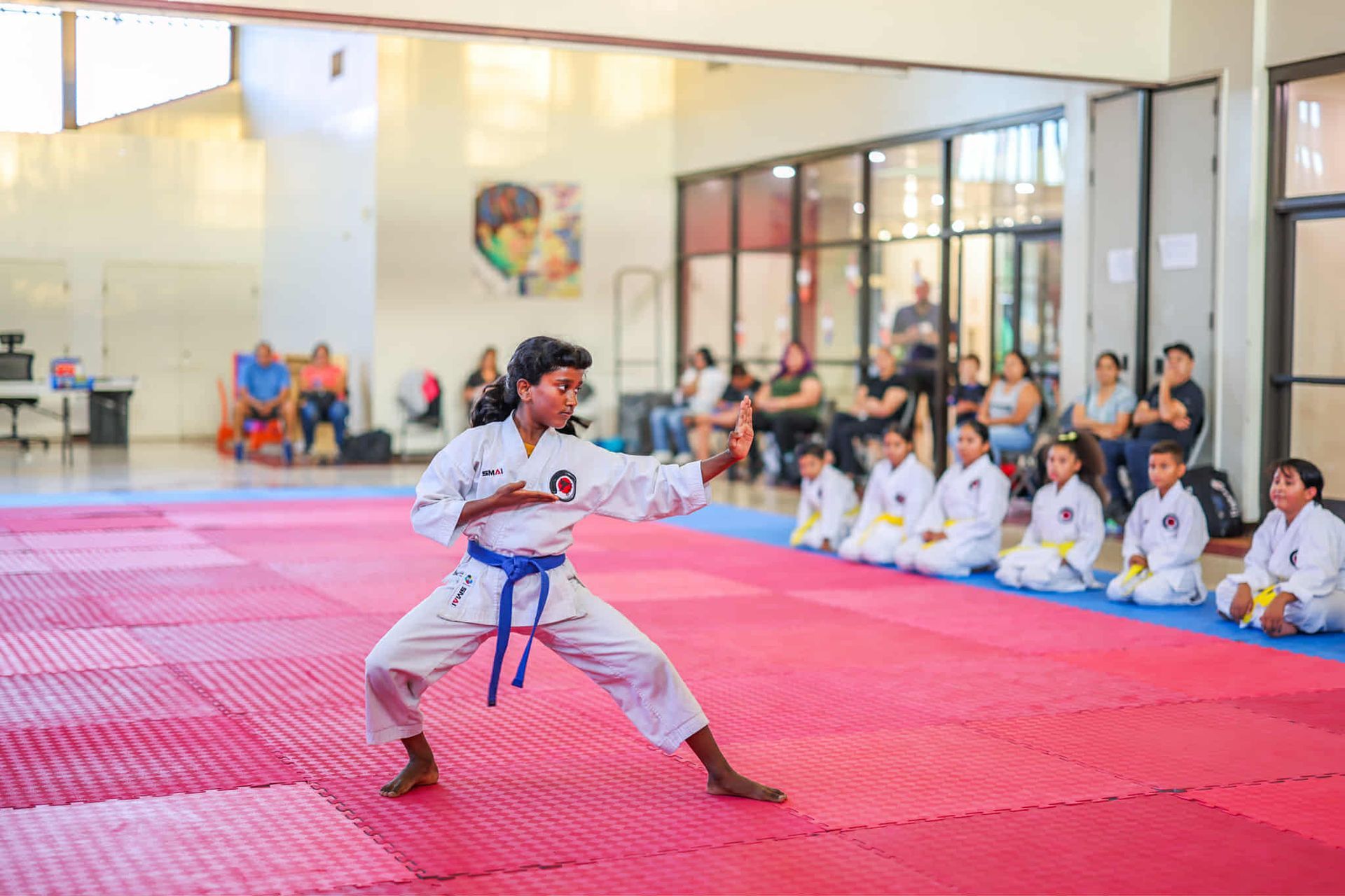 Girl in karate uniform performs kata on a red mat, with other students and spectators in the background.