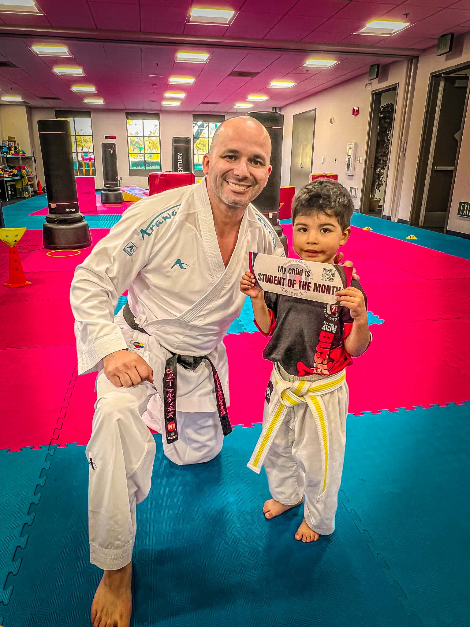 Man in karate uniform kneels, holding certificate with smiling child in karate uniform; dojo background.