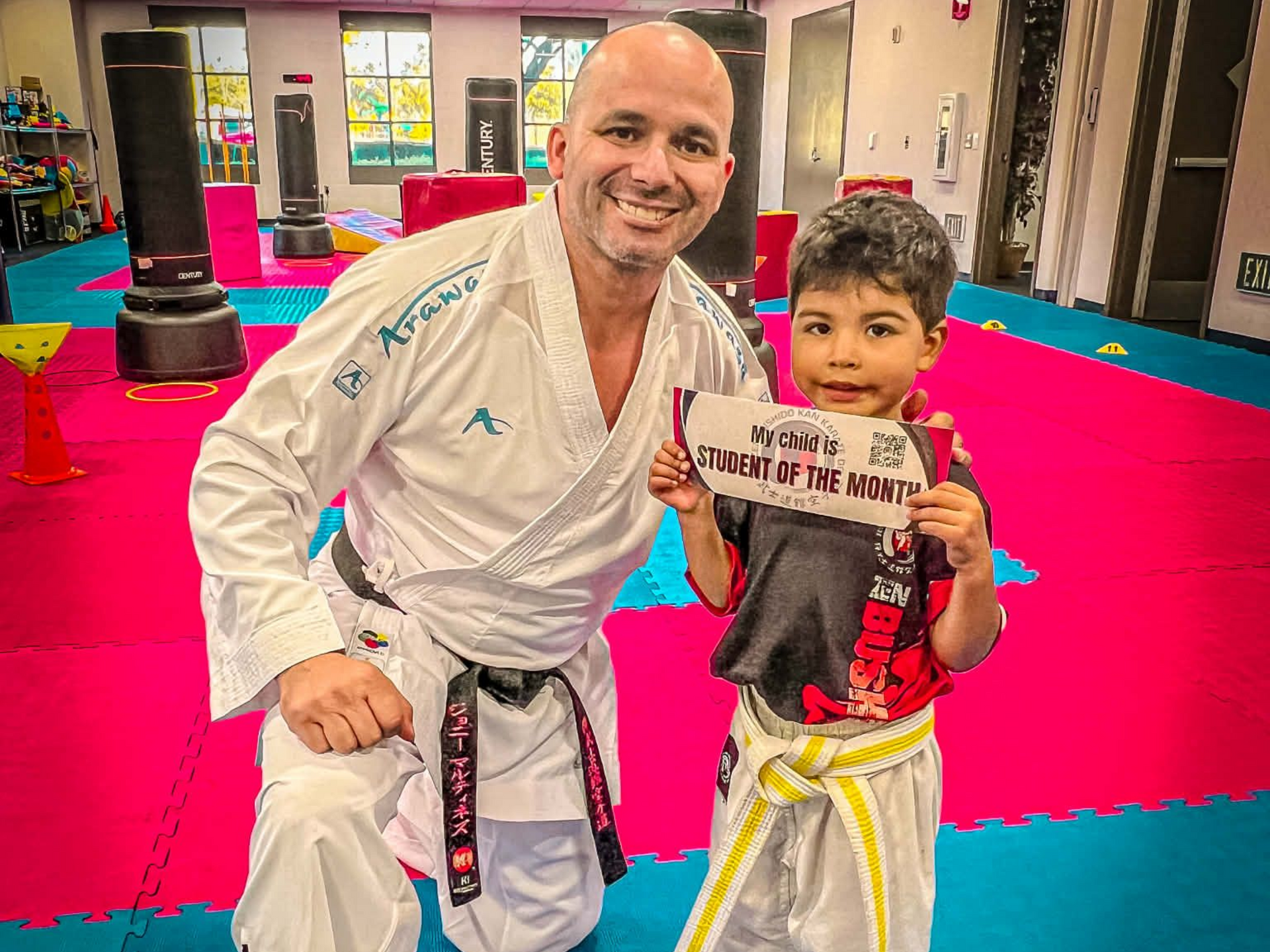 Man in karate uniform kneels, smiling, holding award with young boy in martial arts gear, inside dojo.