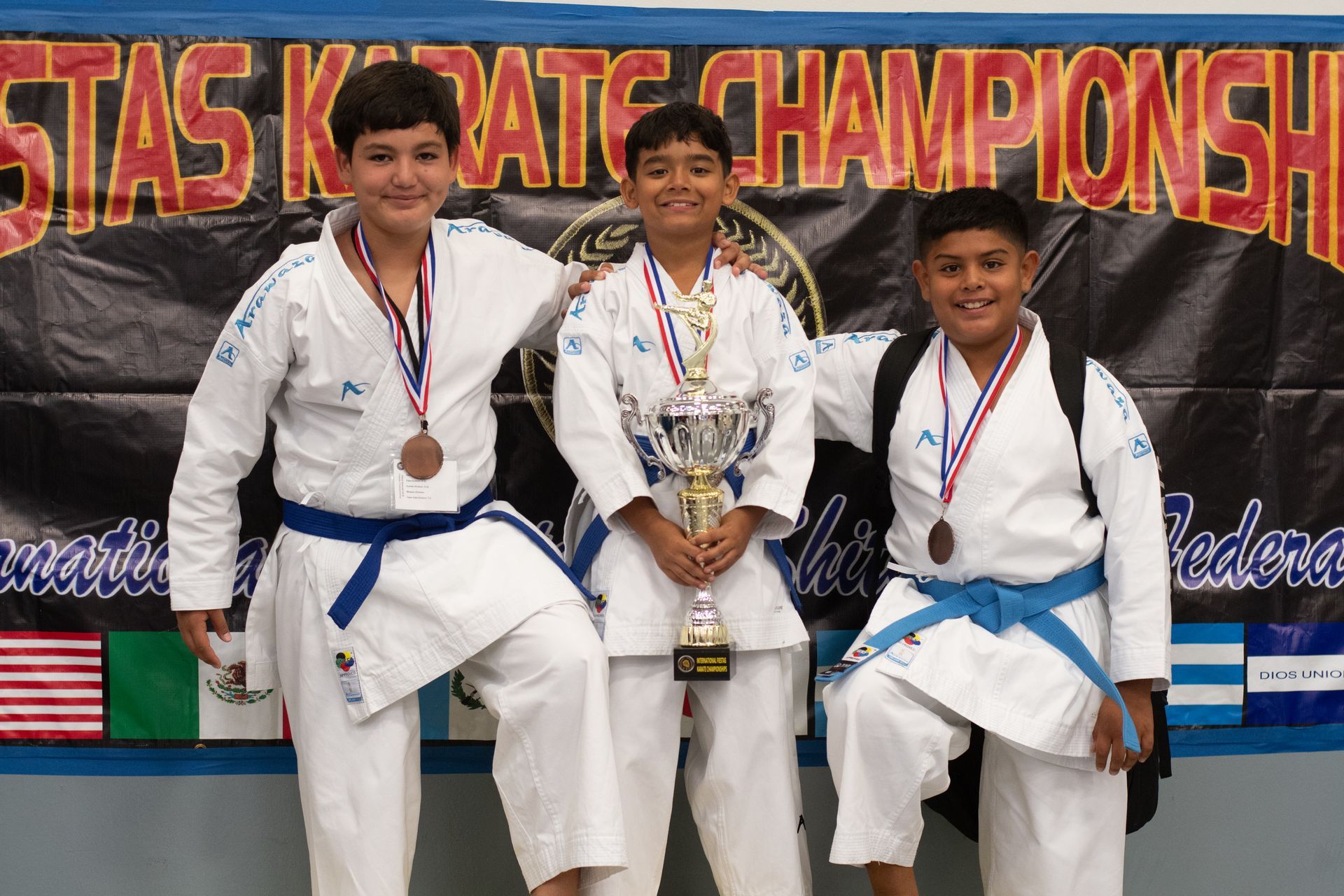 Three boys in karate uniforms at a competition; two with bronze medals, one holding a trophy, and smiling.