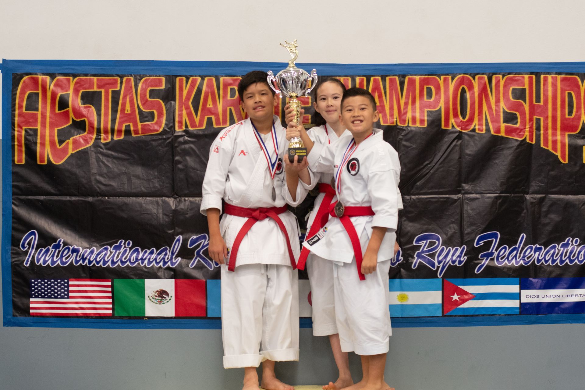 Karate tournament: Three children in white uniforms hold a trophy, winners pose with medals, flags in background.