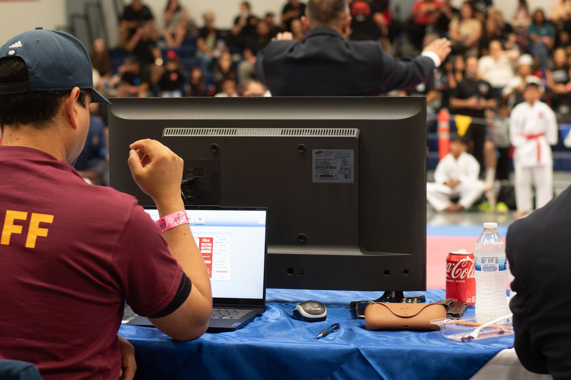 Man at a table with laptop, judging a karate competition. Spectators and a judge in background.