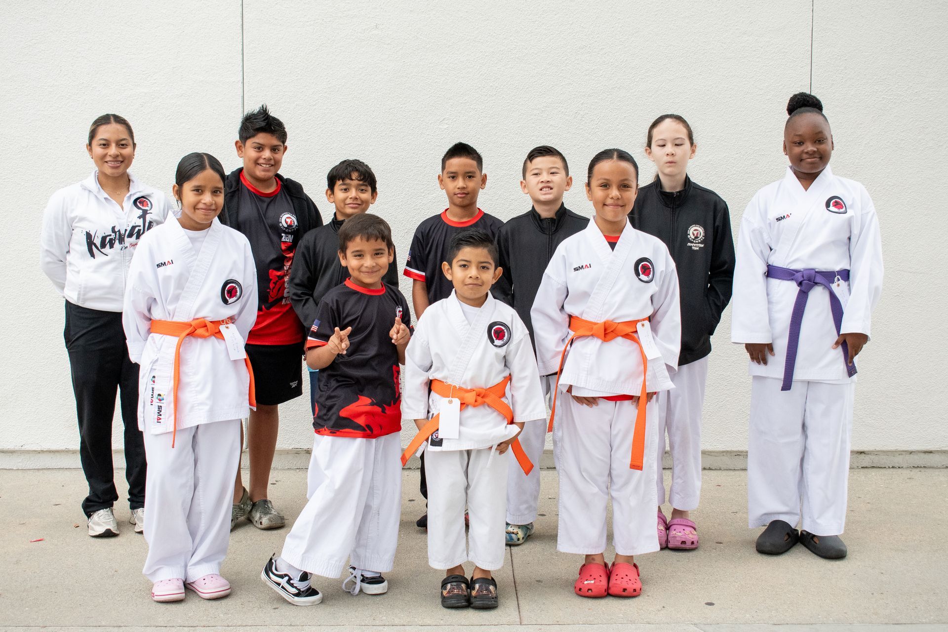 Group of karate students in white gis with colored belts, posing in front of a white wall.
