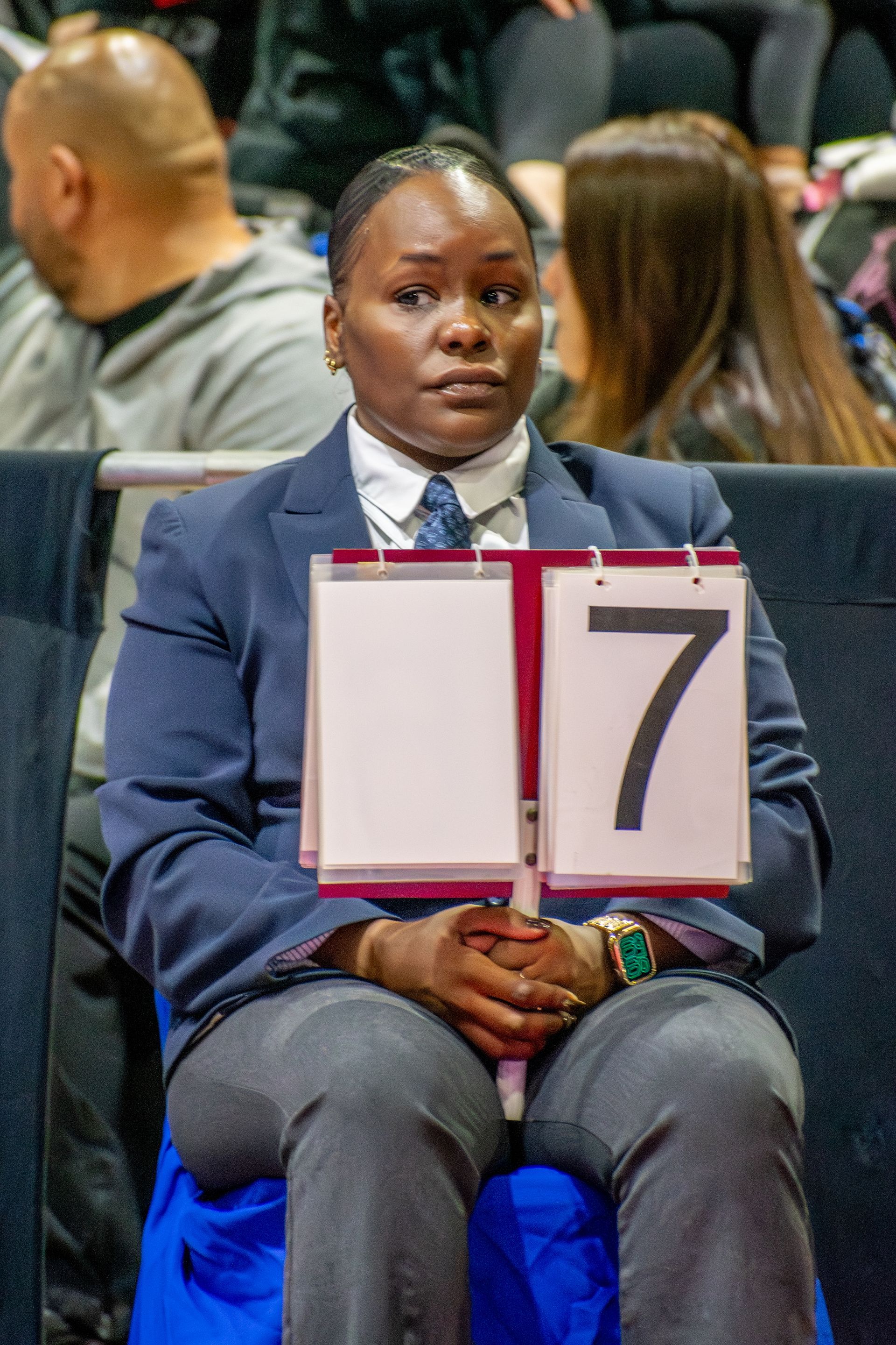 Person in a dark blazer holding a sign with the number 7, sitting in a crowded indoor setting.