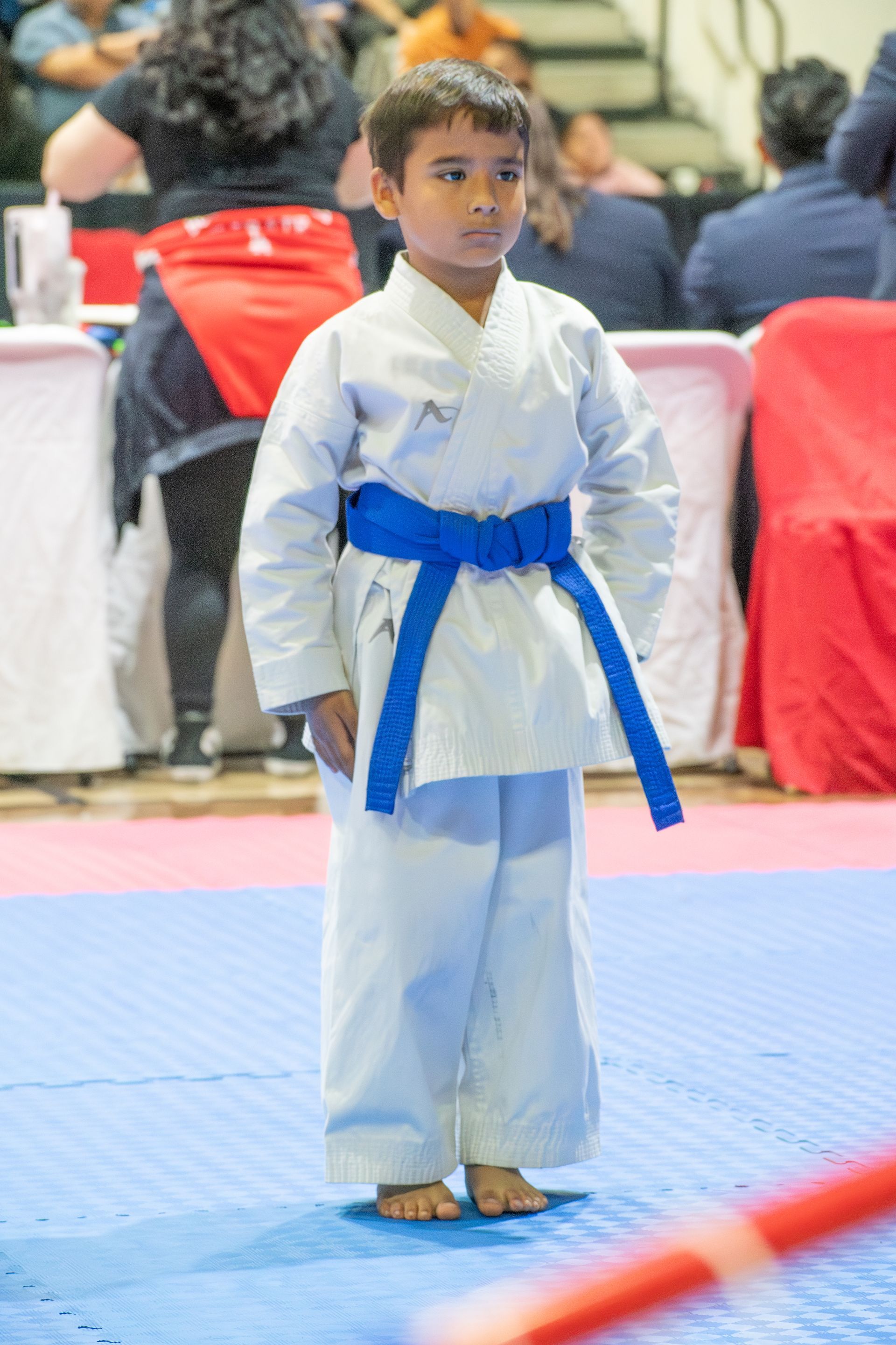 Boy in white karate uniform with blue belt stands on a blue mat.