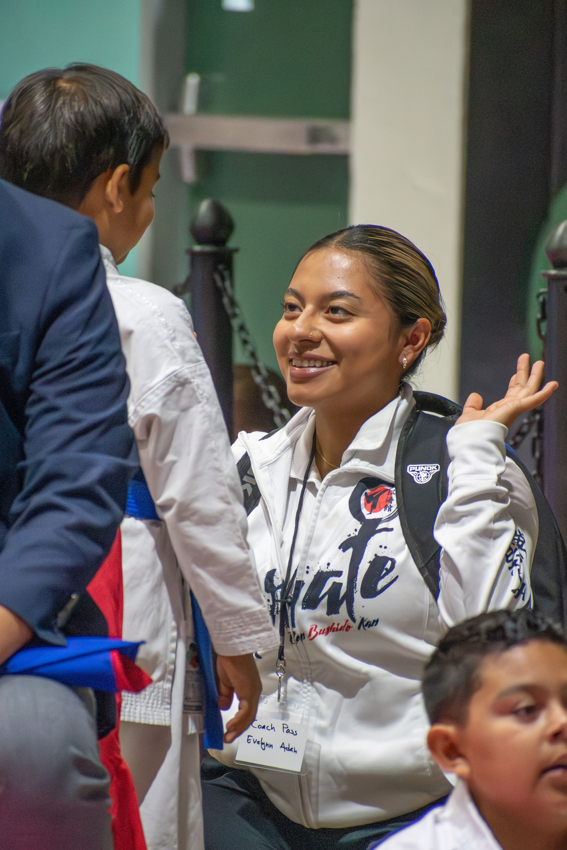 Woman in a karate jacket smiles, talking to a child in a hallway; other children visible.