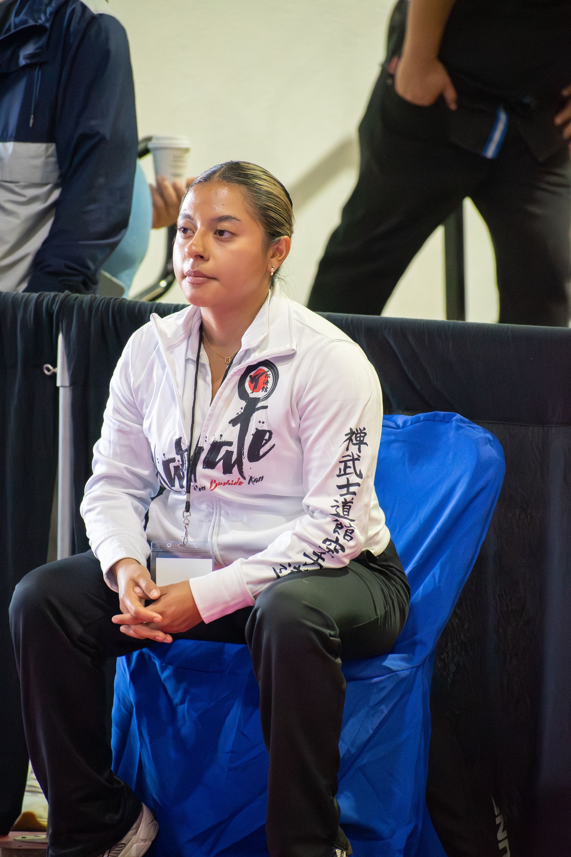 Woman in white Karate jacket seated, looking on attentively.
