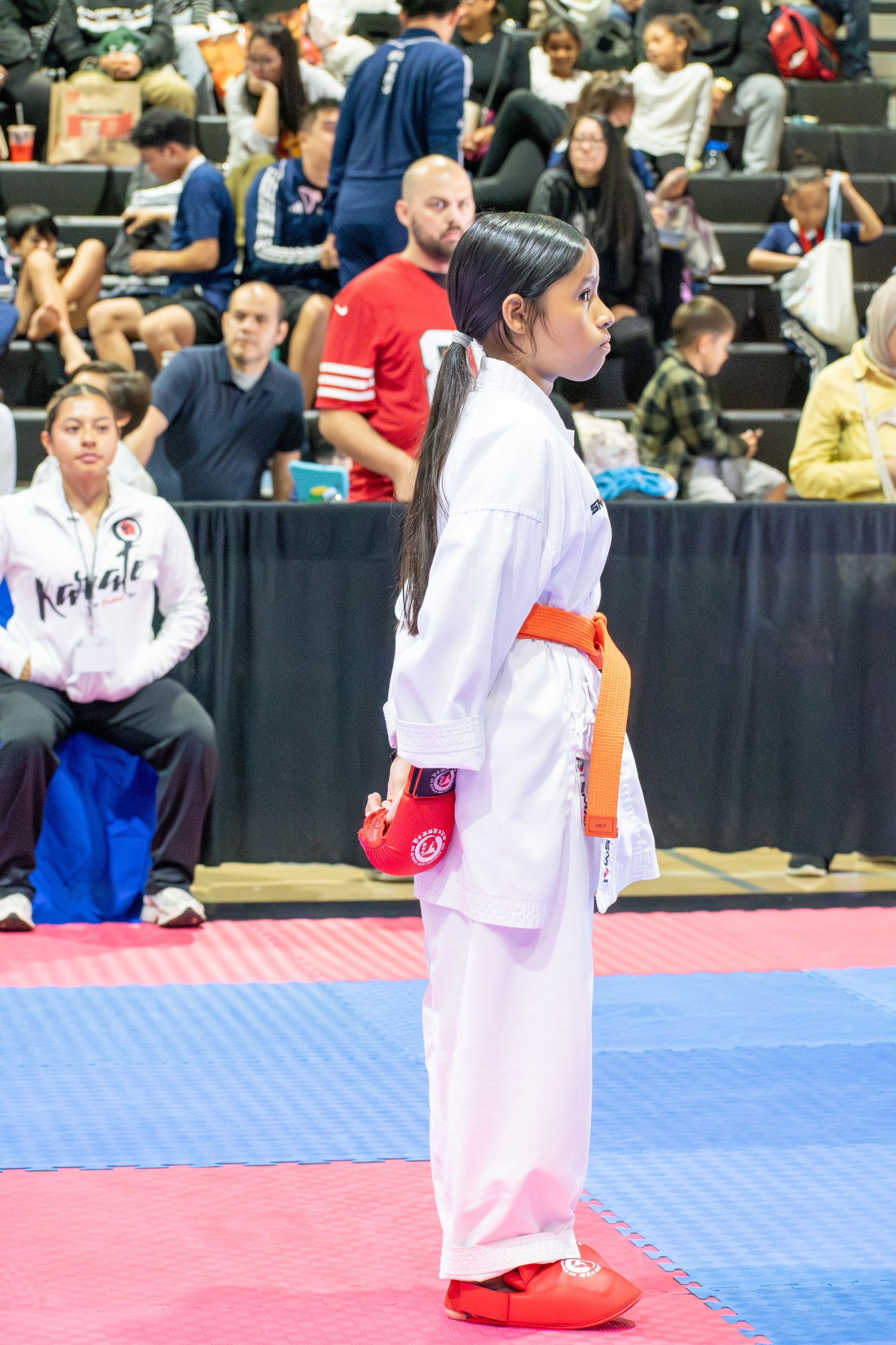 Karate athlete in white uniform with orange belt, red gloves and shoes, on mat, looking ahead.