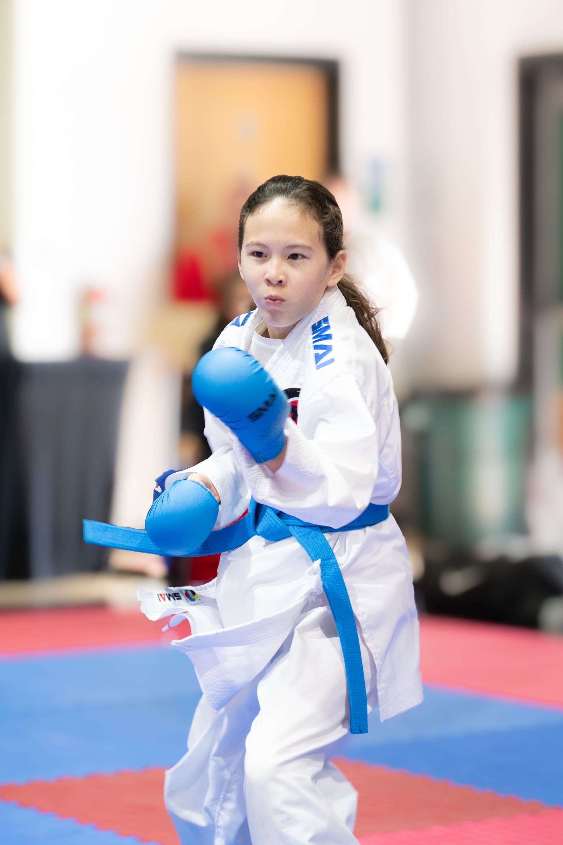 Girl in karate uniform with blue belt and gloves in fighting stance on mat.