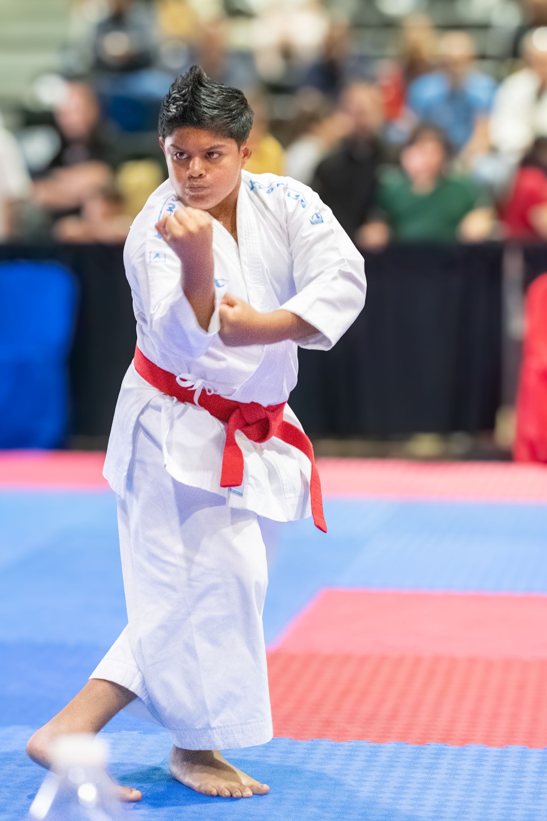 Person in white karate uniform, red belt, in a fighting stance. They are in a competition setting.