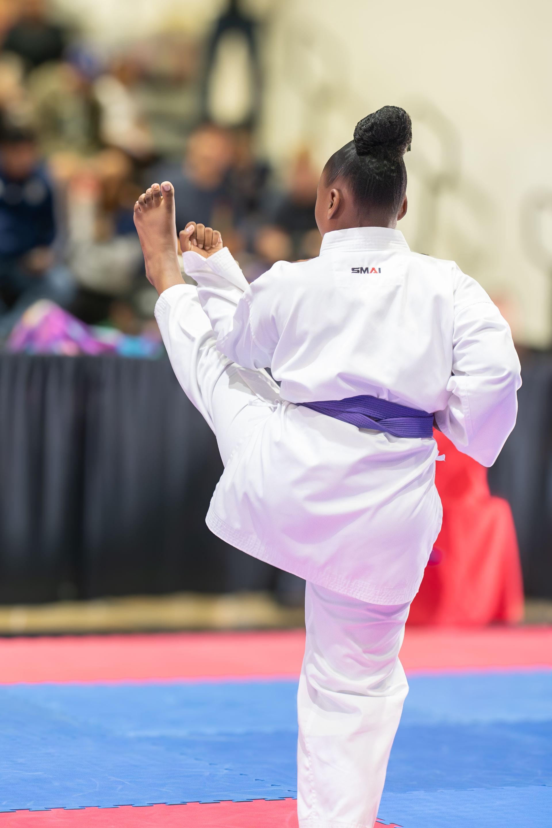 Karateka in white uniform kicks during a competition, purple belt, indoor setting.