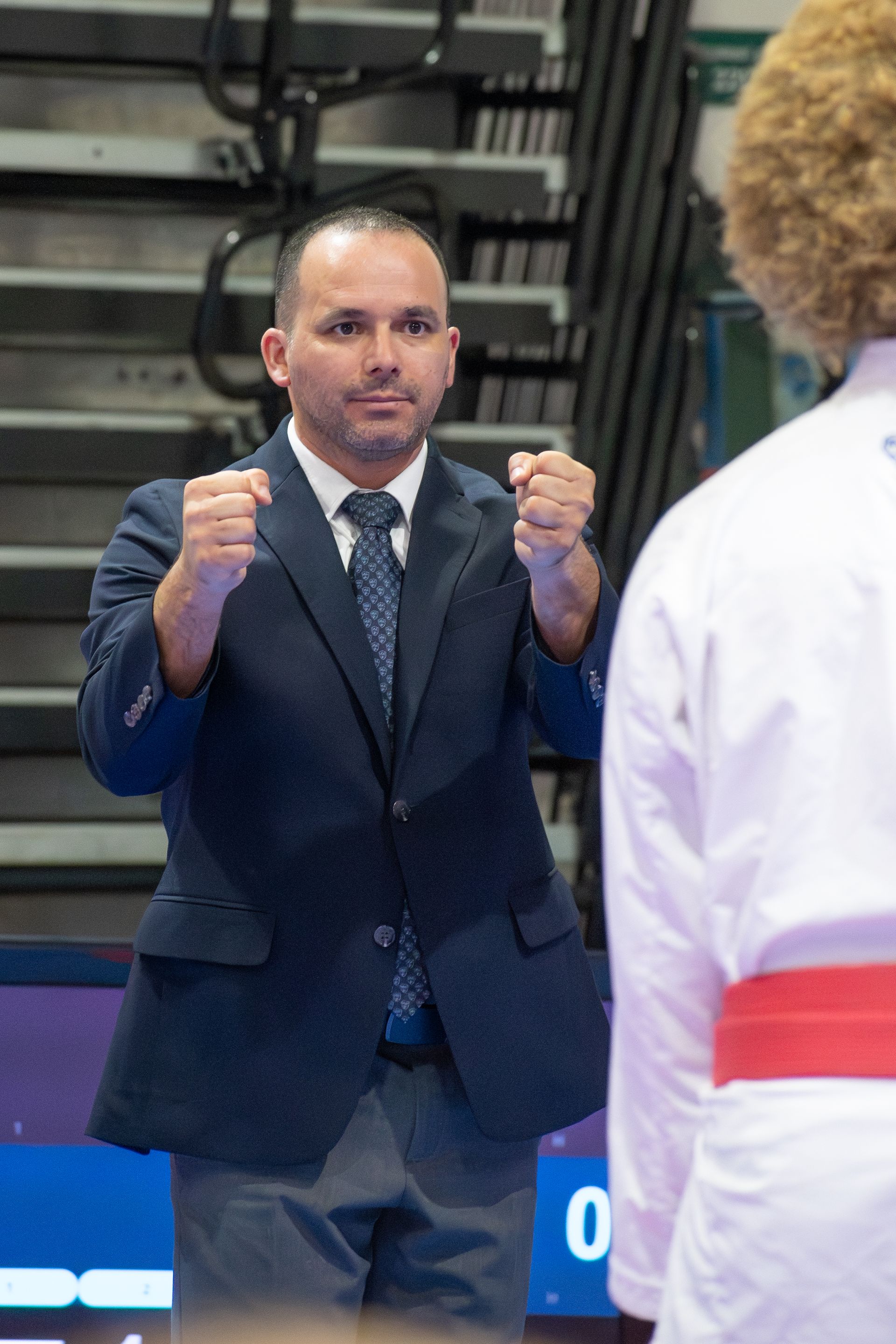 Man in suit with fists up, facing person in karate uniform at competition.