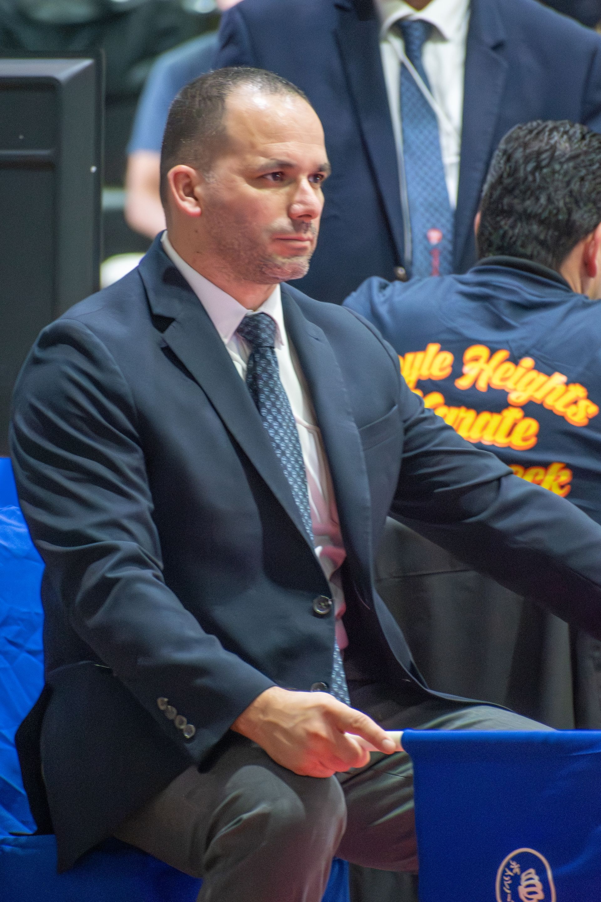 Man in a navy blazer sits, looking focused, at a sporting event.