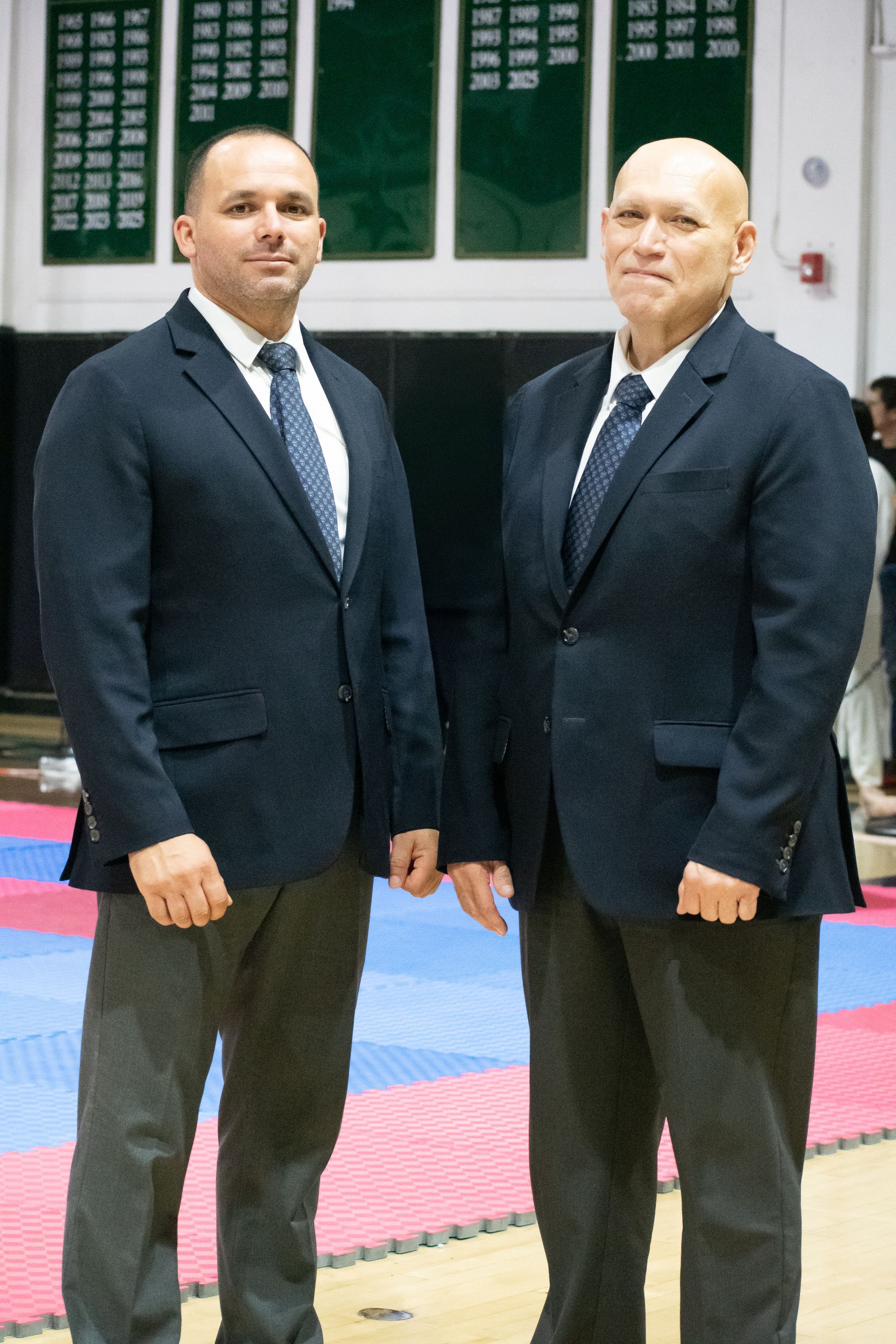 Two men in blazers and ties standing side by side in a gymnasium setting. They are in front of what appears to be a competition mat.