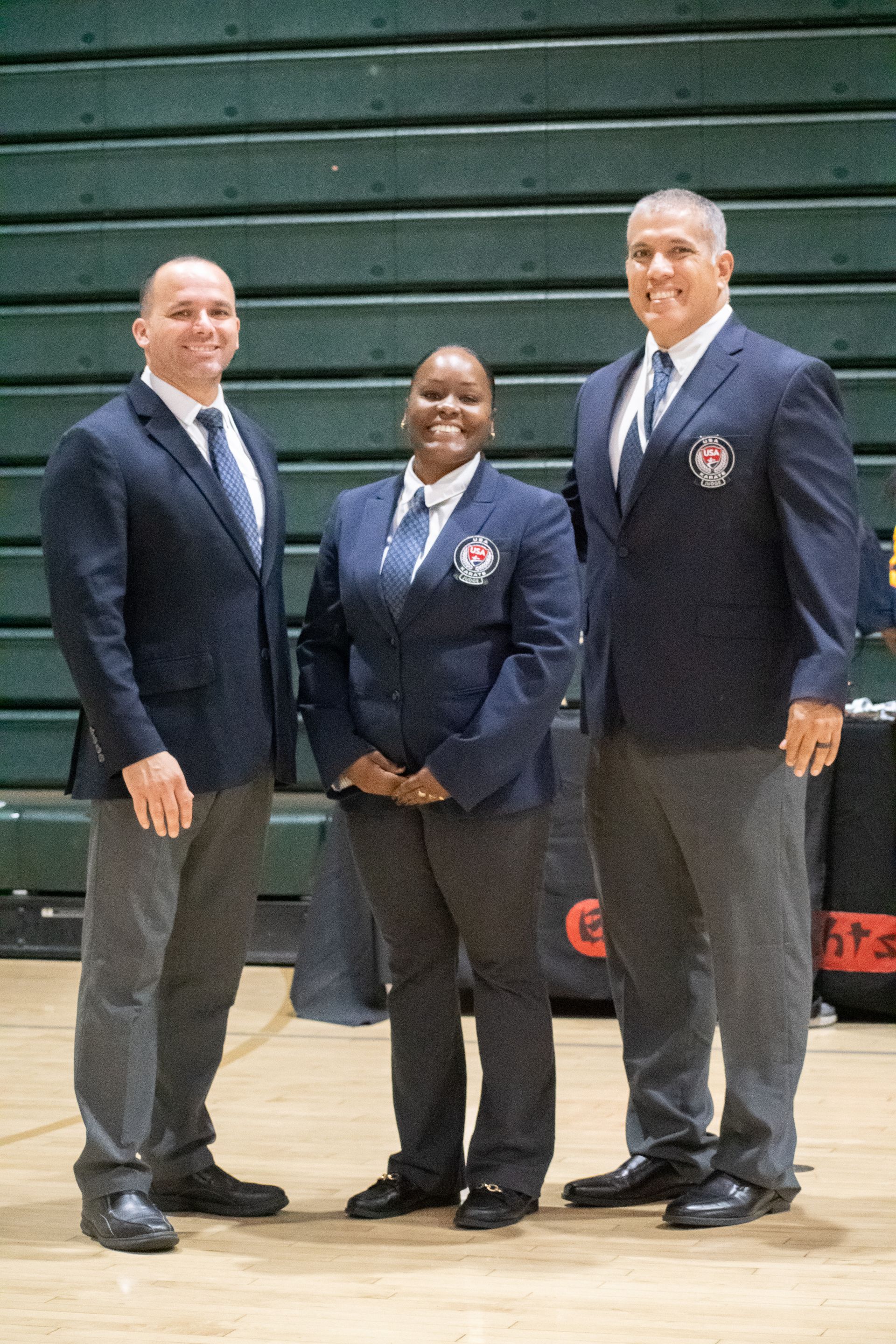 Three people in navy blazers, white shirts, ties. Center person smiles. Green background.