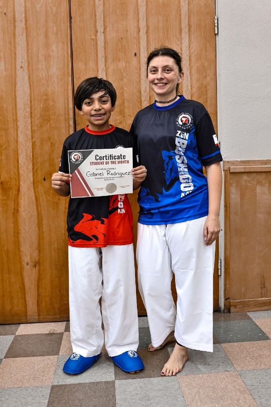 Two youths holding a certificate, standing indoors in martial arts uniforms against wooden doors.